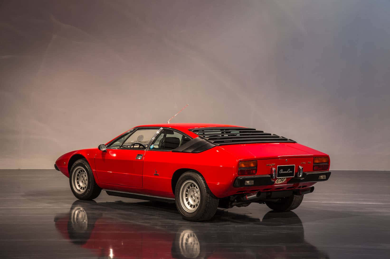 Lamborghini Urraco 桌布 壁紙 — 一輛充滿活力的紅色經典跑車，設計流暢，帶有黑色點綴，在柔和的工作室燈光下展示在反光地板上。