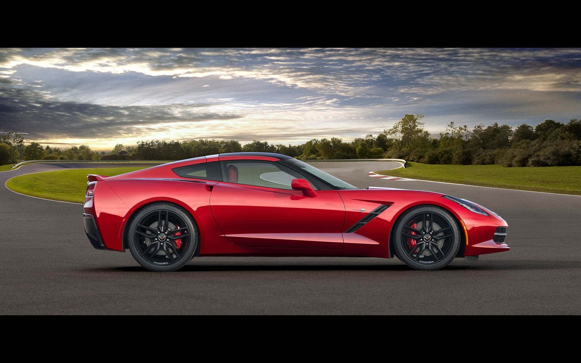Chevrolet Corvette Grand Sport 桌布 壁紙 — 一輛充滿活力的紅色跑車在日落時分戲劇性的多雲天空下沿著蜿蜒的瀝青賽道滑行。