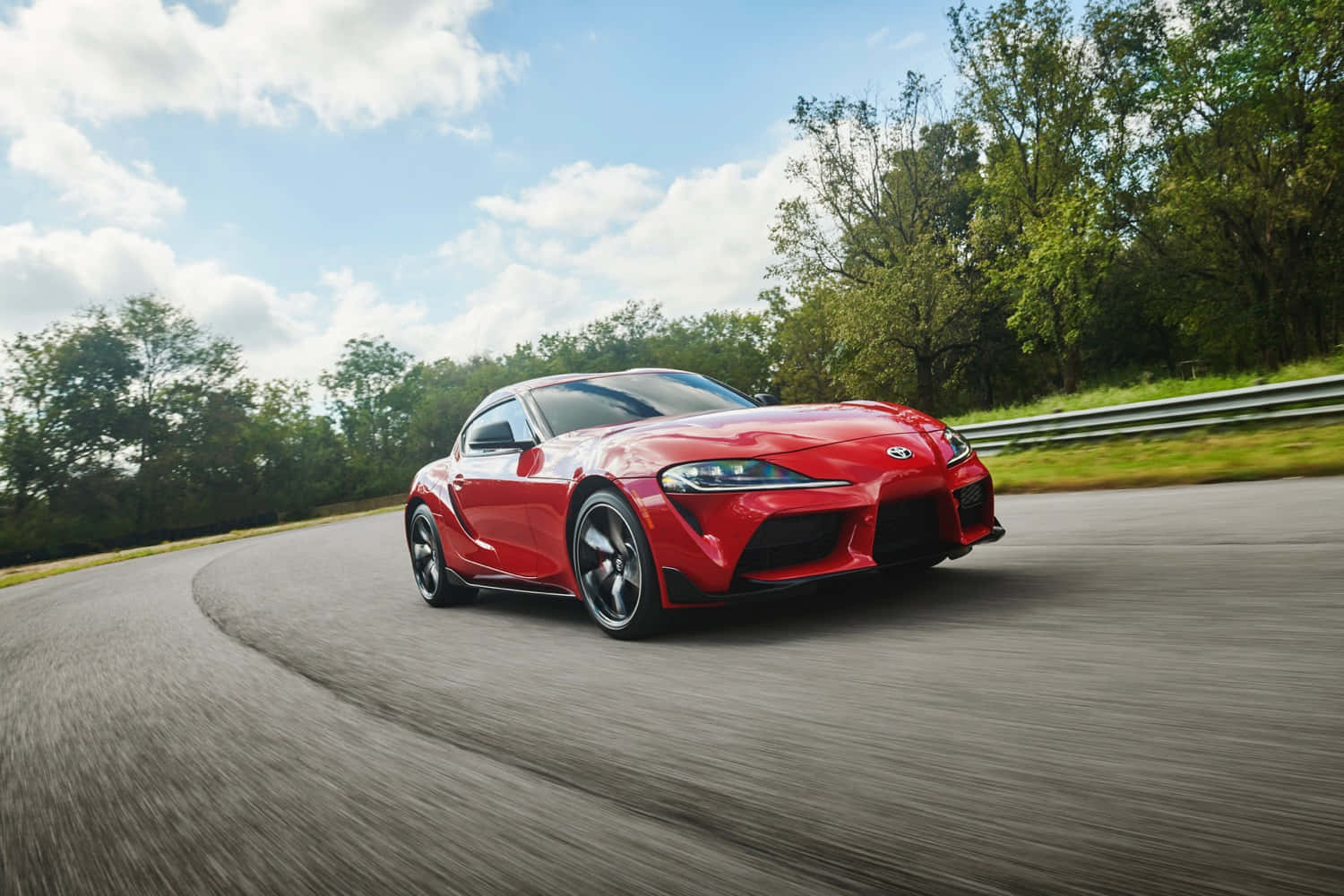 Best Car 桌布 壁紙 — 一輛充滿活力的紅色跑車沿著一條彎曲的瀝青道路疾馳，周圍環繞著茂密的綠樹，天空湛藍，白雲朵朵。