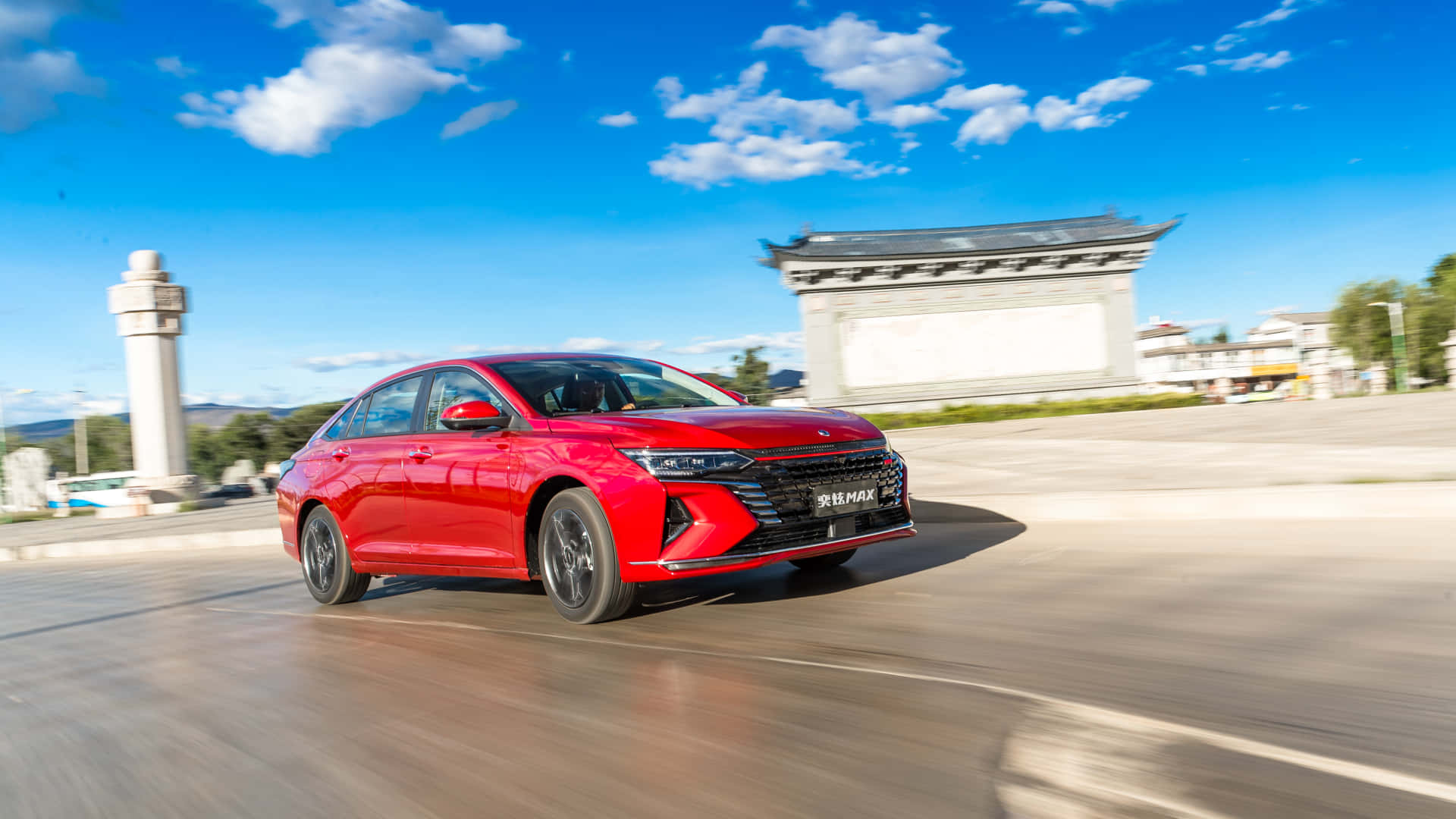 Dongfeng 桌布 壁紙 — 一輛充滿活力的紅色轎車在傳統建築附近，於明亮的藍天和蓬鬆的白雲下行駛在鋪砌的道路上。