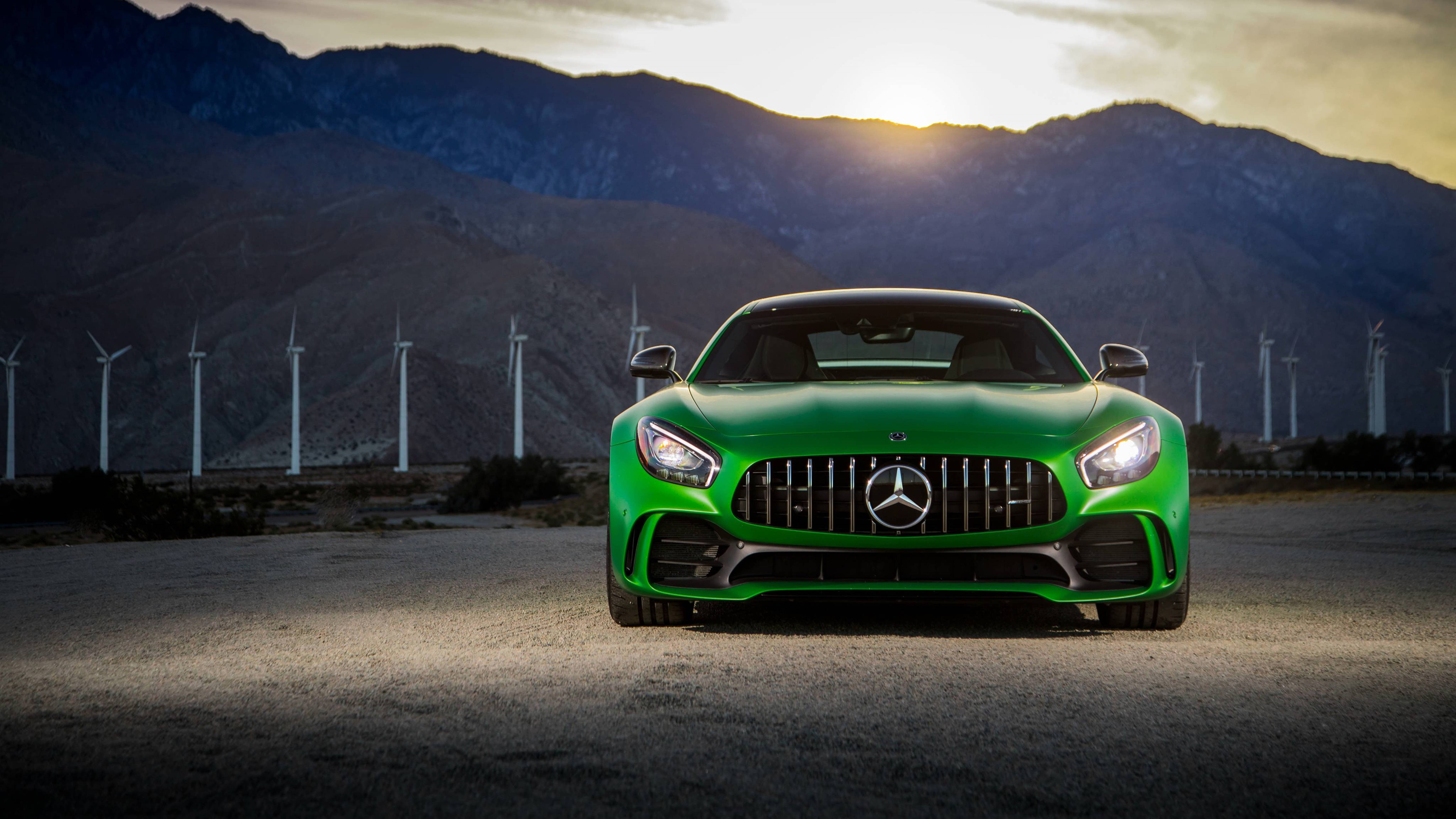 Mercedes 桌布 壁紙 — 一輛充滿活力的綠色 Mercedes-AMG GT 跑車在沙漠公路上向前行駛，背景有風力發電機和山脈，天空是金色的日落。