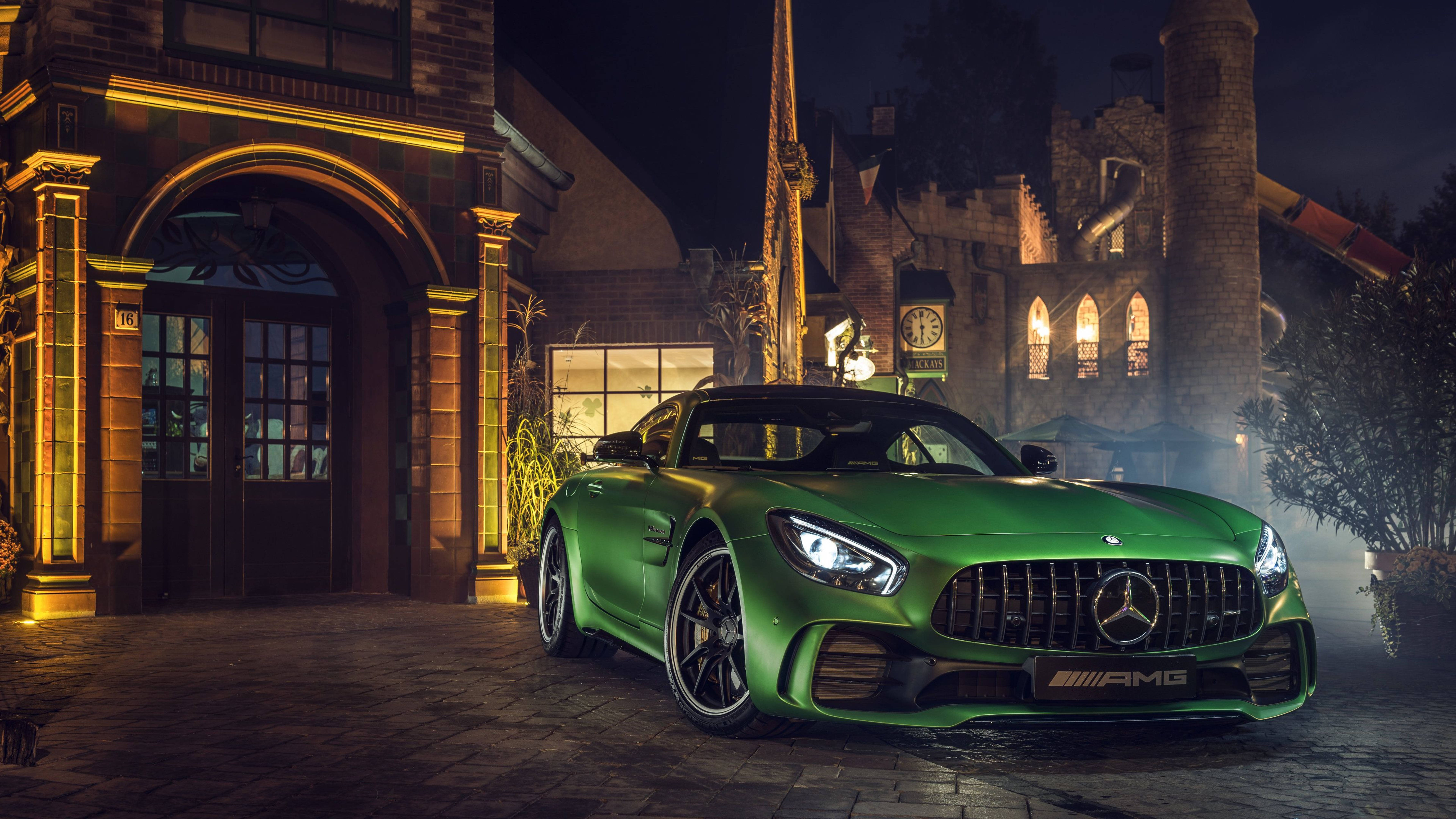 平治 AMG 桌布 壁紙 — 一輛充滿活力的綠色 Mercedes-AMG GT 跑車在溫暖的夜燈下，於一座宏偉的石建築前閃閃發光，建築物窗戶透出光芒，氣氛迷濛。
