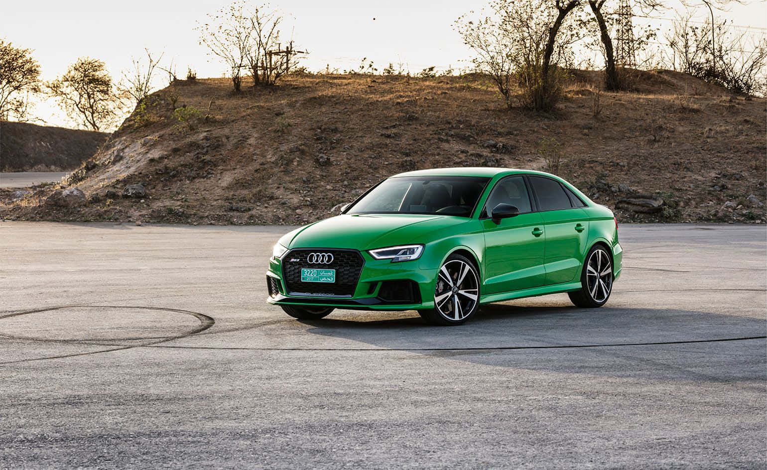 Audi Rs3 桌布 壁紙 — 一輛充滿活力的綠色Audi RS3轎車停在帶有輪胎痕的混凝土停車場上，背景是乾燥的山坡，正值黃金時刻。