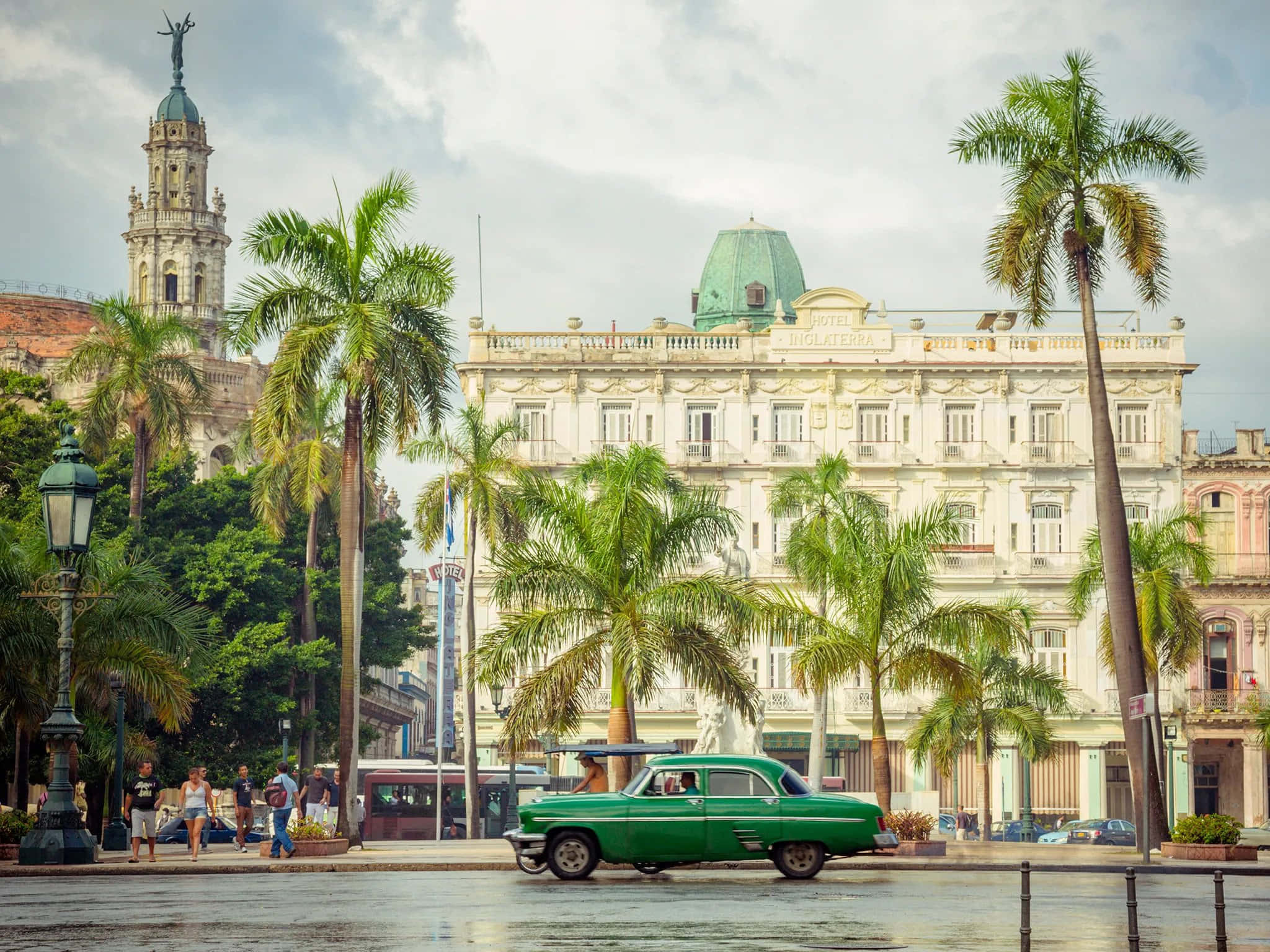 Cuba 桌布 壁紙 — 一輛充滿活力的綠色復古汽車在哈瓦那多雲的天空下行駛，經過棕櫚樹和殖民時期建築。