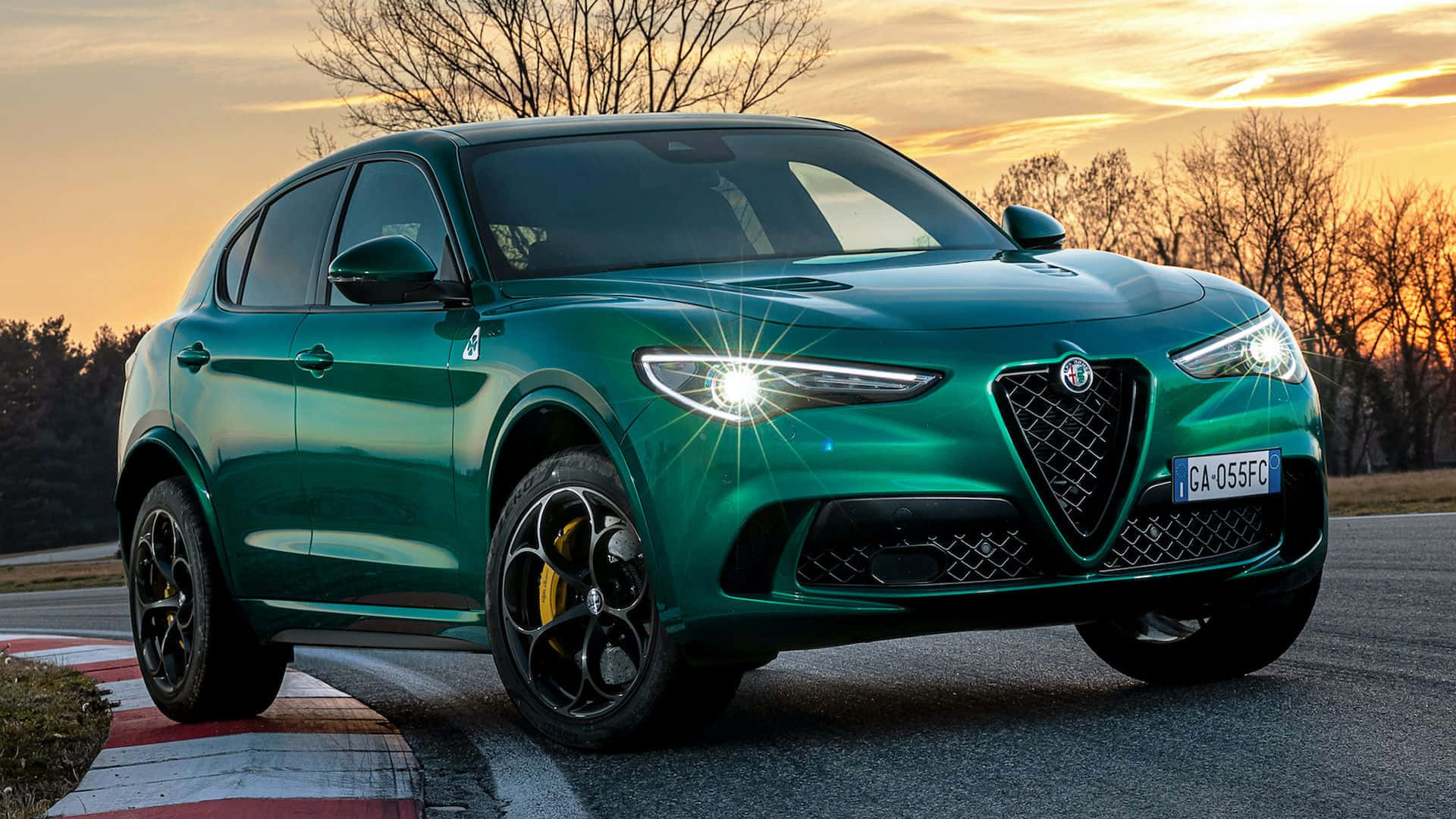 Alfa Romeo Stelvio 桌布 壁紙 — 一輛充滿活力的綠色愛快羅密歐SUV在日落時分的賽道上閃閃發光，背景是光禿禿的樹木。