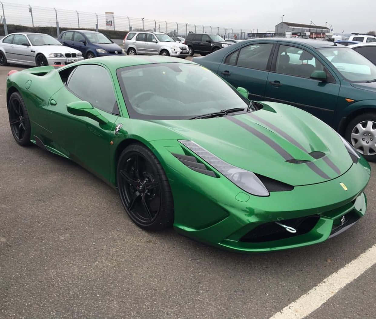 Ferrari 458 Speciale 桌布 壁紙 — 一輛充滿活力的綠色法拉利跑車帶有黑色賽車條紋，停放在停車場中，與其他車輛一起，天空多雲。