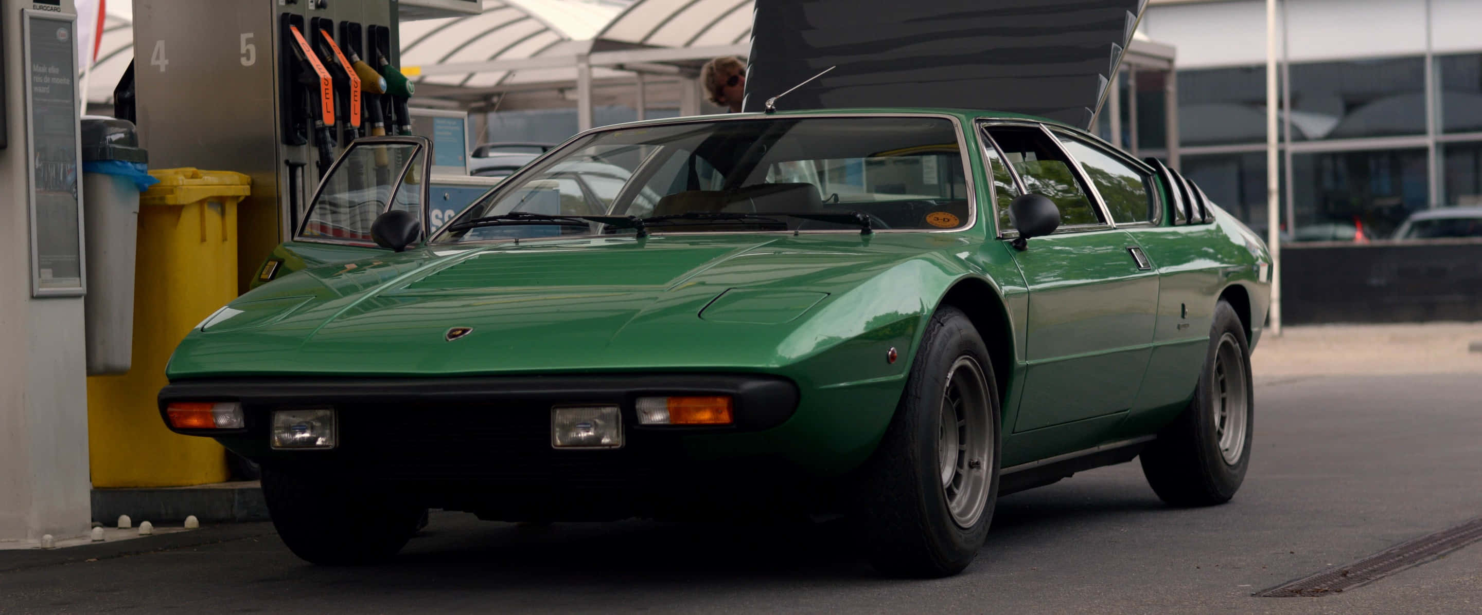 Lamborghini Urraco 桌布 壁紙 — 一輛充滿活力的綠色經典跑車打開引擎蓋，停放在日光下的現代加油站。