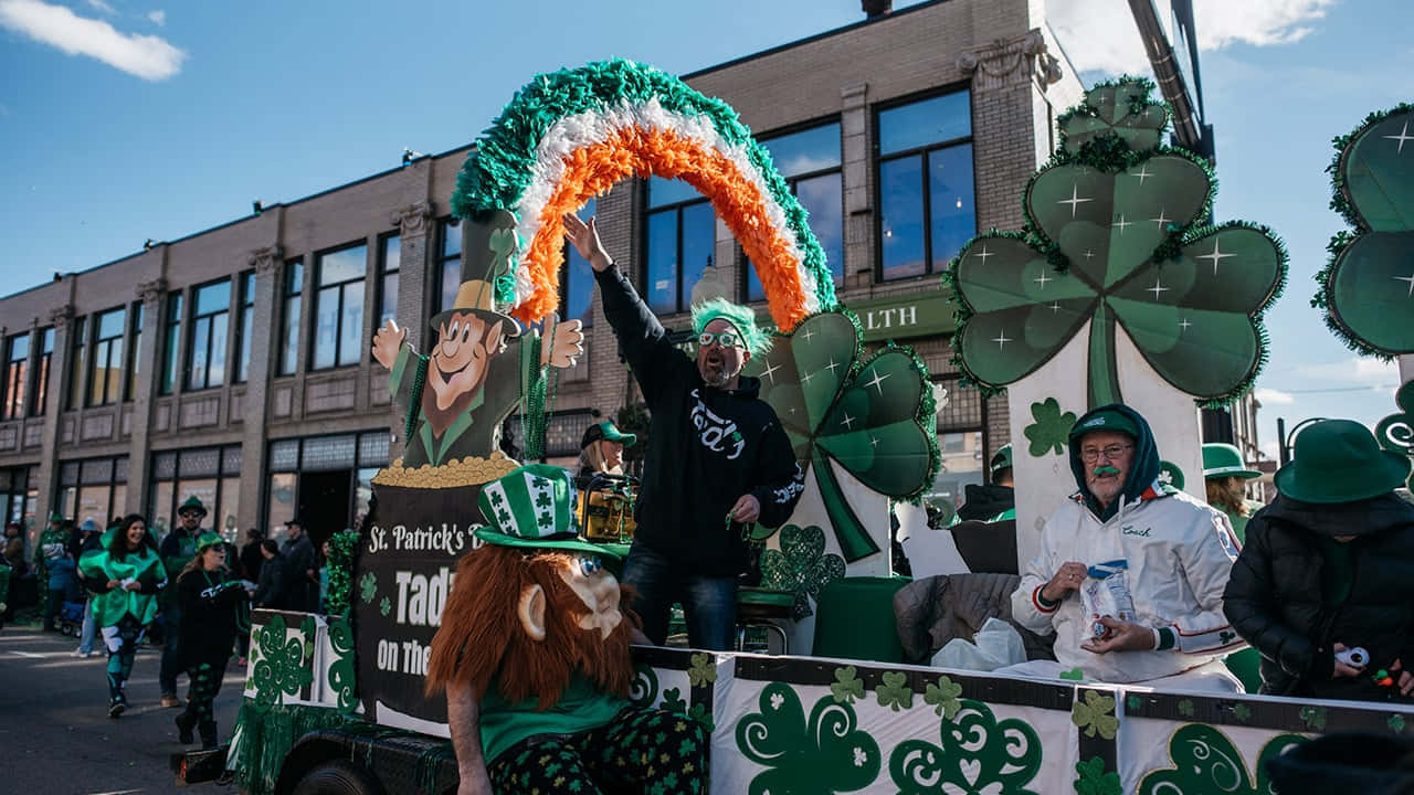St Patrick's Day 桌布 壁紙 — 一輛充滿活力的聖帕特里克節巡遊花車，裝飾著綠色三葉草和一道彩虹，拱形在穿著節日服裝的歡呼狂歡者上方。