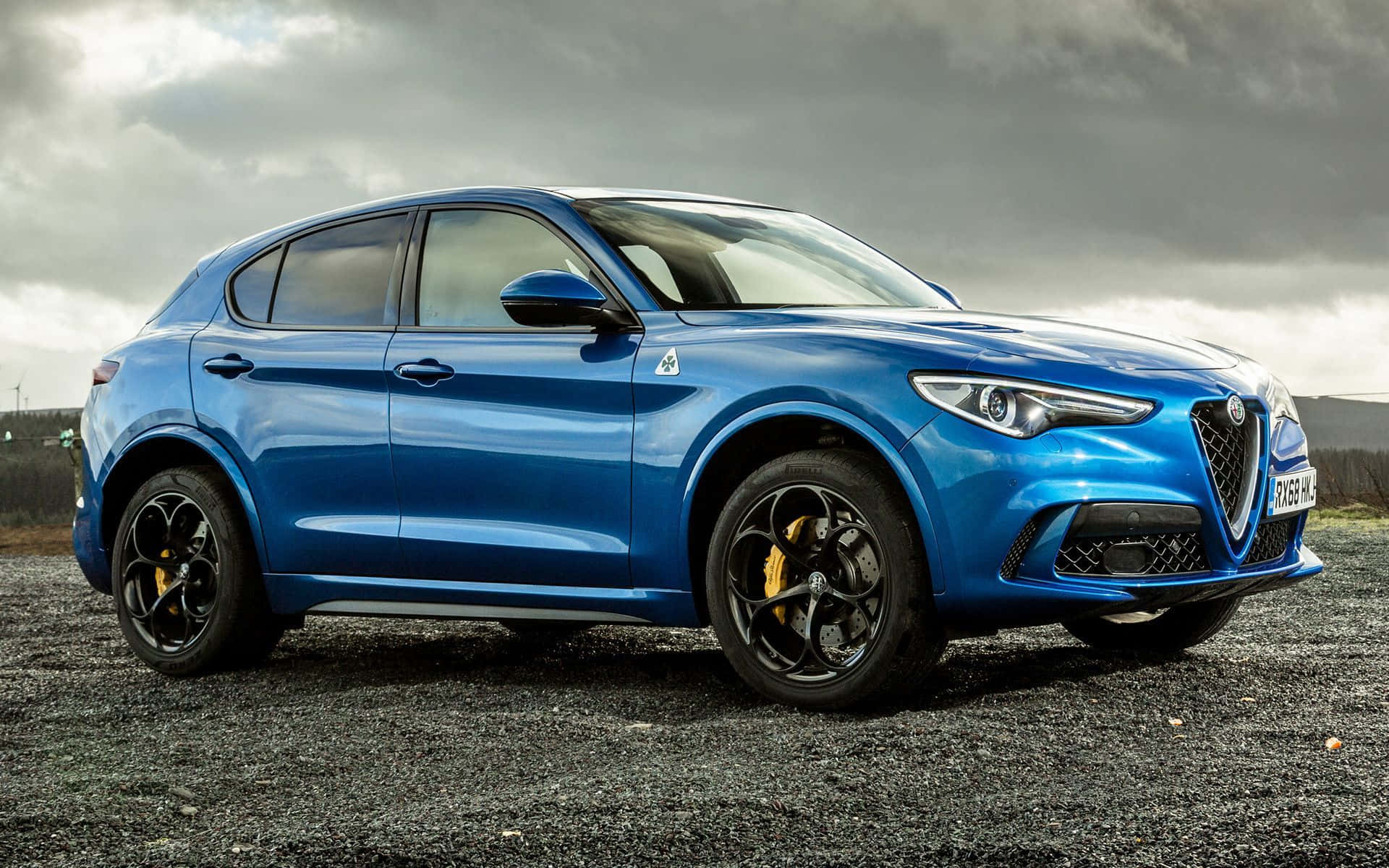 Alfa Romeo Stelvio 桌布 壁紙 — 一輛充滿活力的藍色 Alfa Romeo SUV，配備黑色車輪和黃色制動卡鉗，停泊在碎石地上，天空多雲。