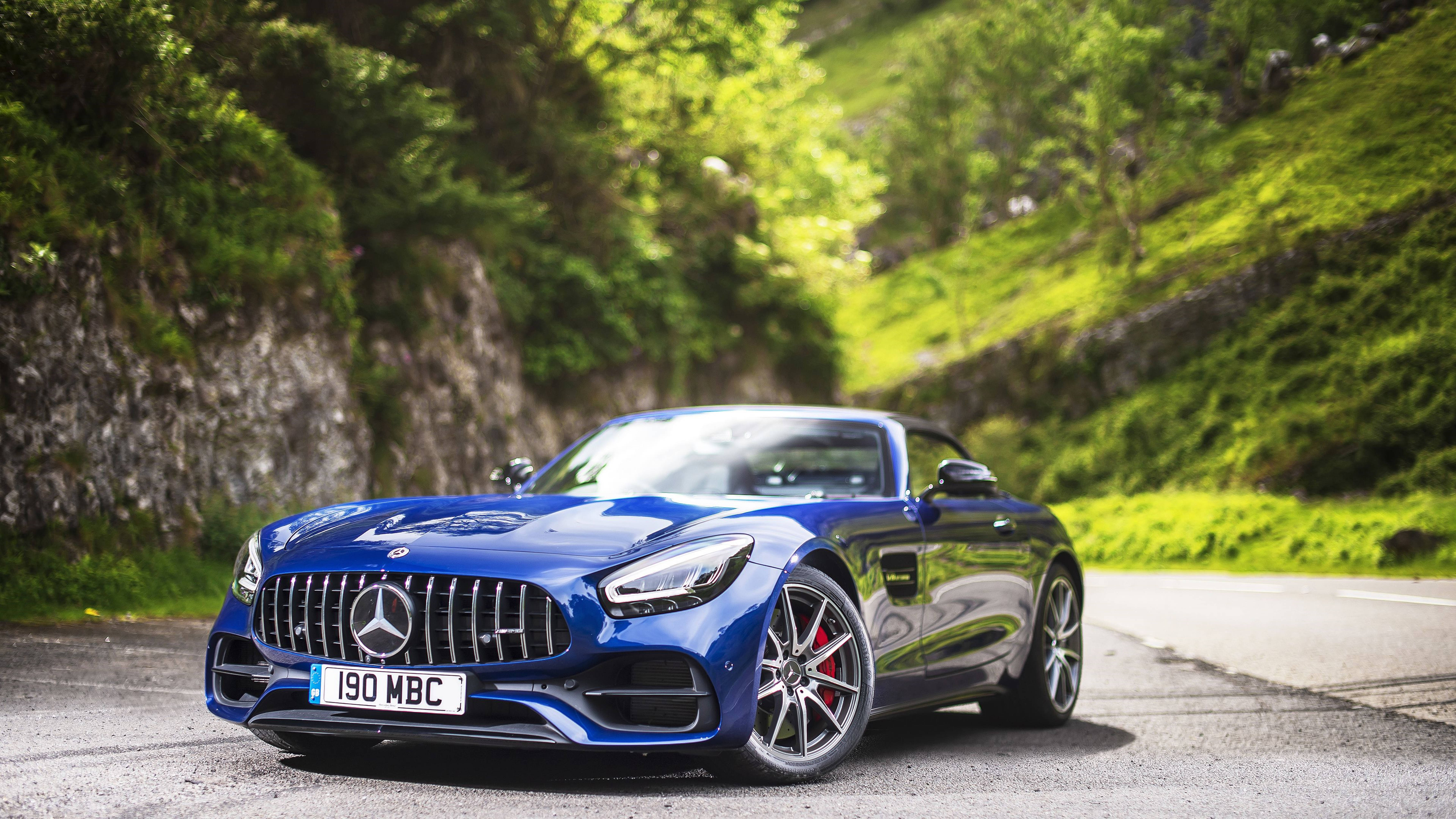 藍色 桌布 壁紙 — 一輛充滿活力的藍色 Mercedes-AMG GT Roadster 在蜿蜒的山路上閃閃發光，周圍環繞著茂密的綠色樹葉。