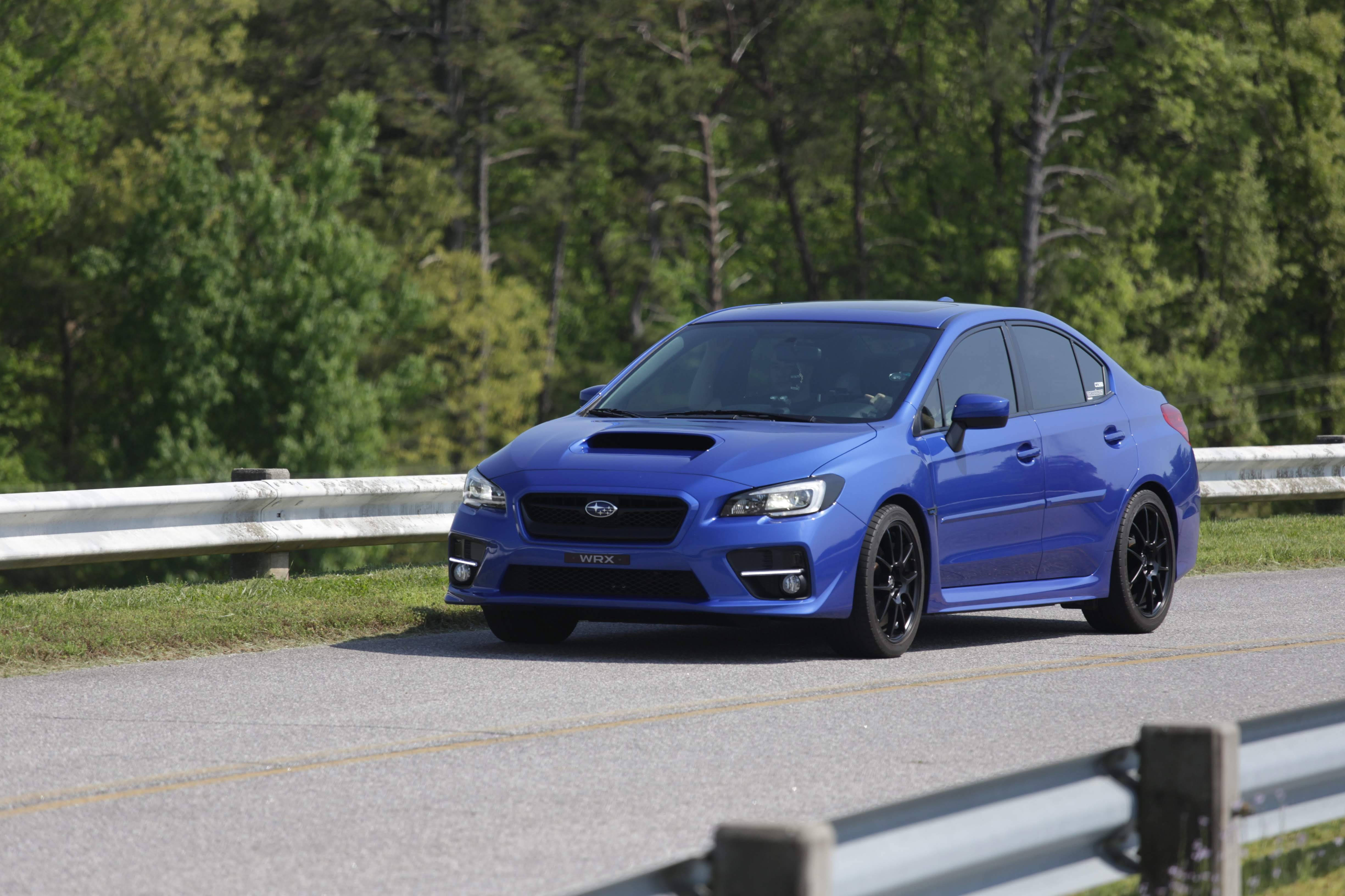 subaru 桌布 壁紙 — 一輛充滿活力的藍色 Subaru WRX 房車在明亮的日光下行駛在一條有護欄的綠樹成蔭的道路上。