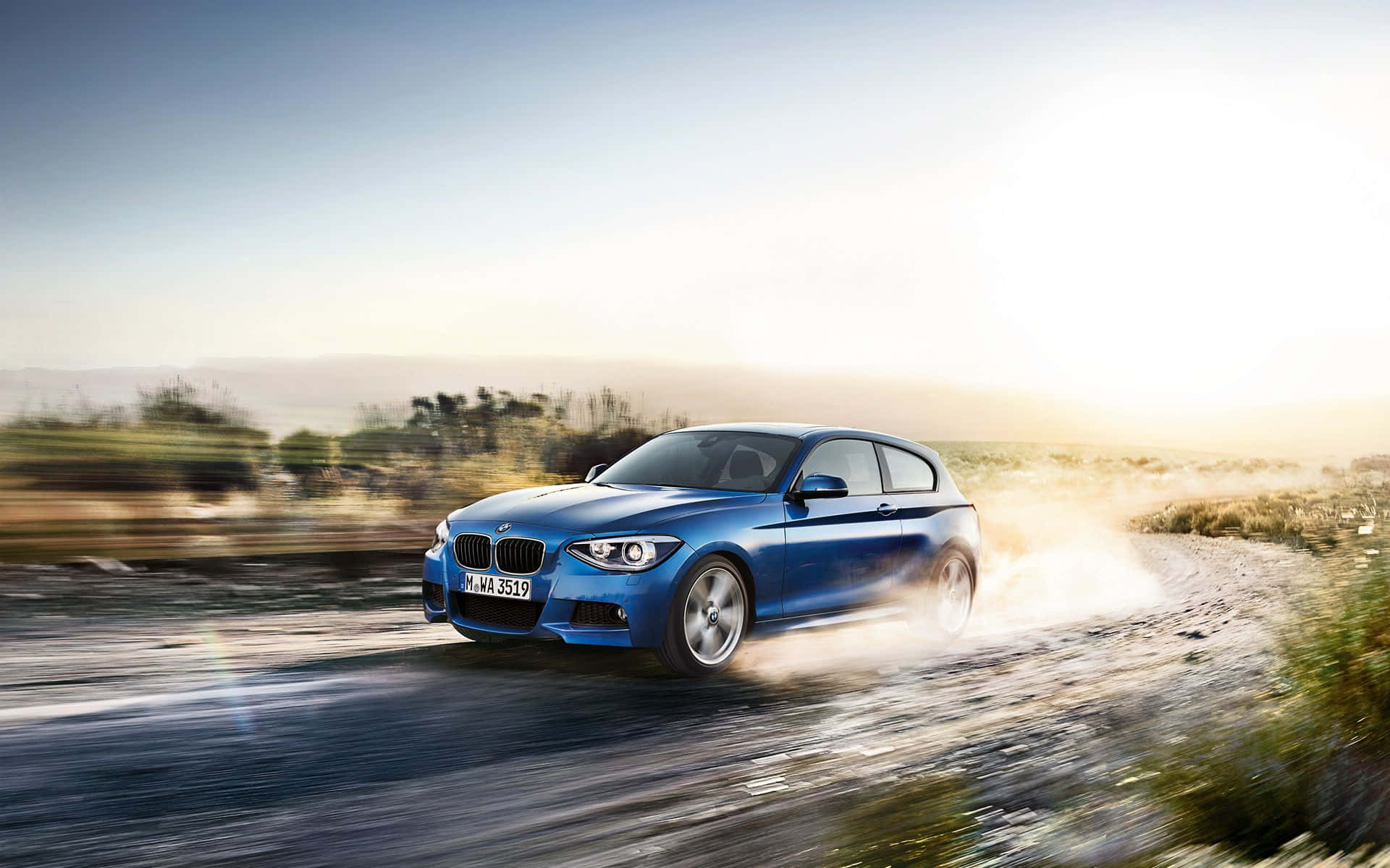 Bmw 1 Series 桌布 壁紙 — 一輛充滿活力的藍色BMW 1系在塵土飛揚的鄉村道路上疾馳，在明亮的金色天空下揚起碎石。