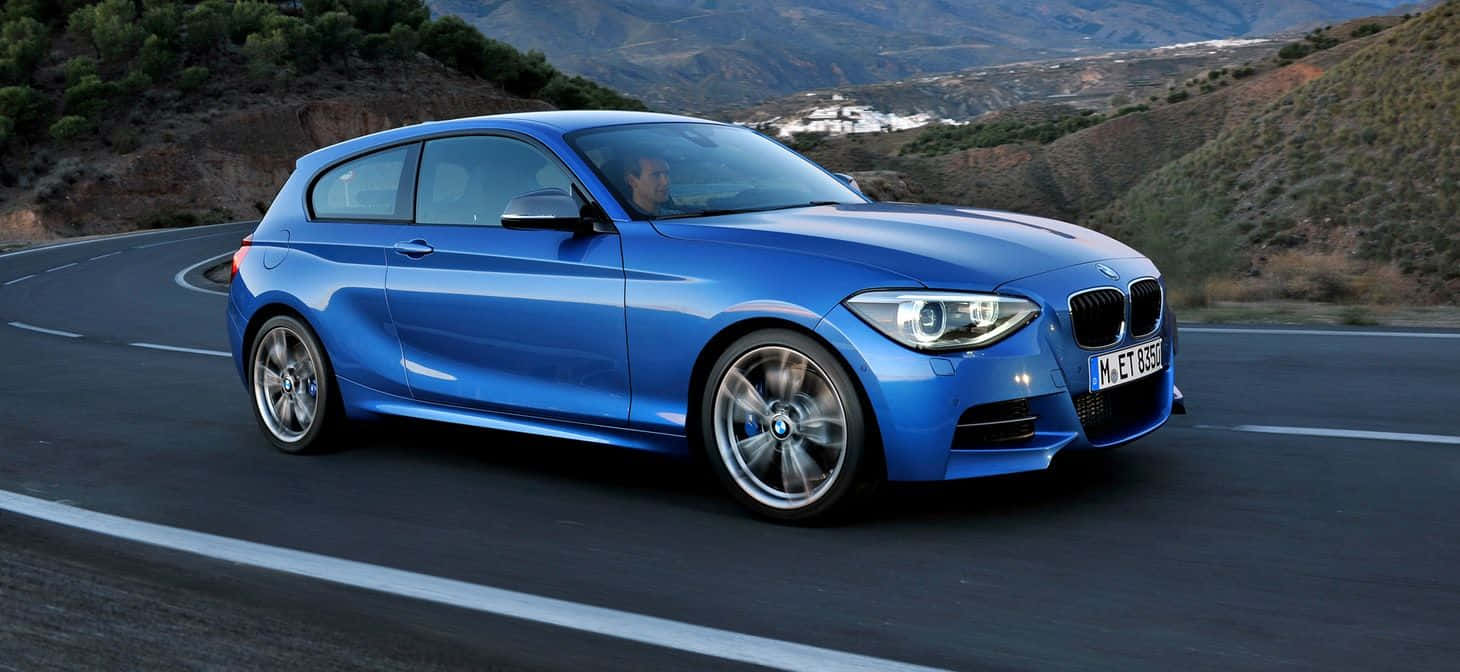 Bmw 1 Series 桌布 壁紙 — 一輛充滿活力的藍色BMW 1系掀背車沿著蜿蜒的山路飛馳，背景是崎嶇的山丘和遠處的村莊。