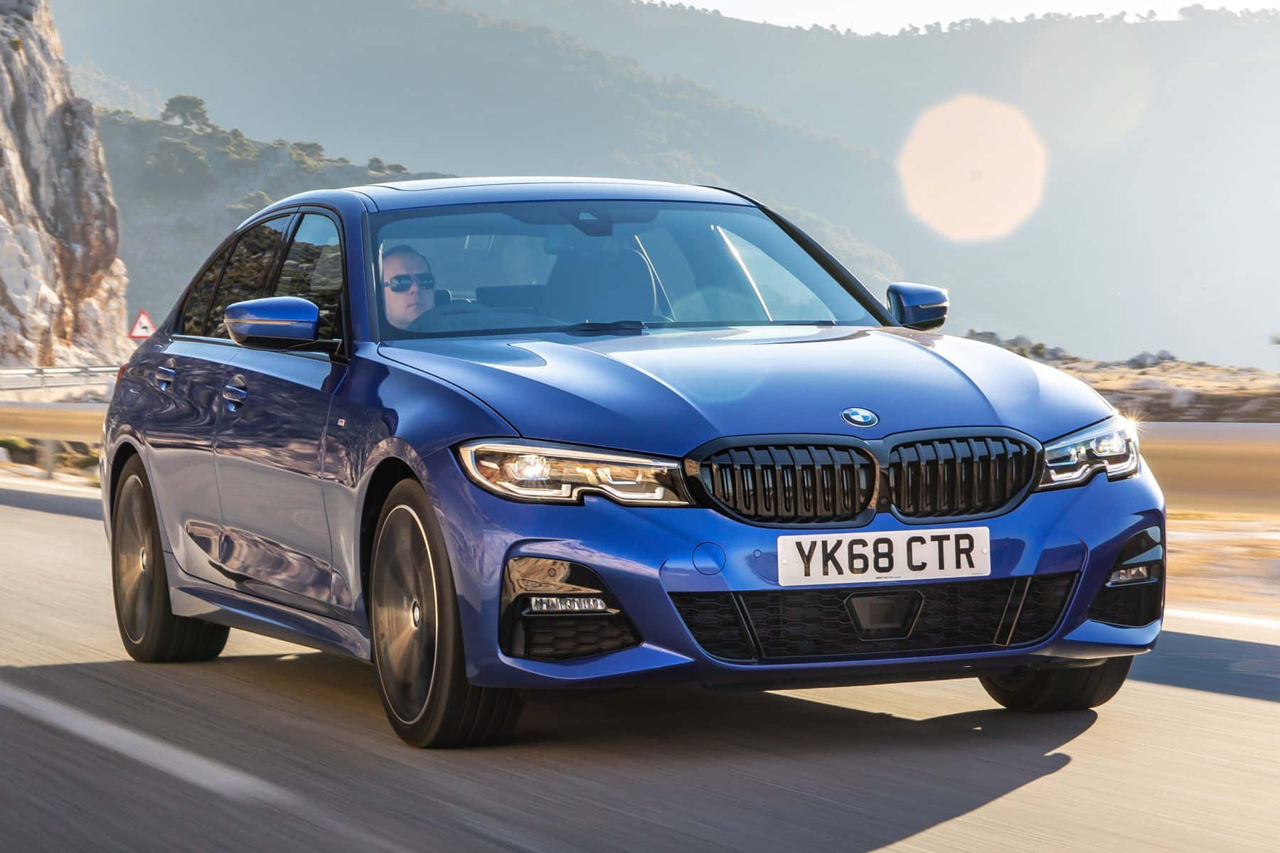 Bmw 3 Series 桌布 壁紙 — 一輛充滿活力的藍色BMW 3系轎車沿著海岸公路飛馳，背景有太陽耀斑和岩石峭壁。