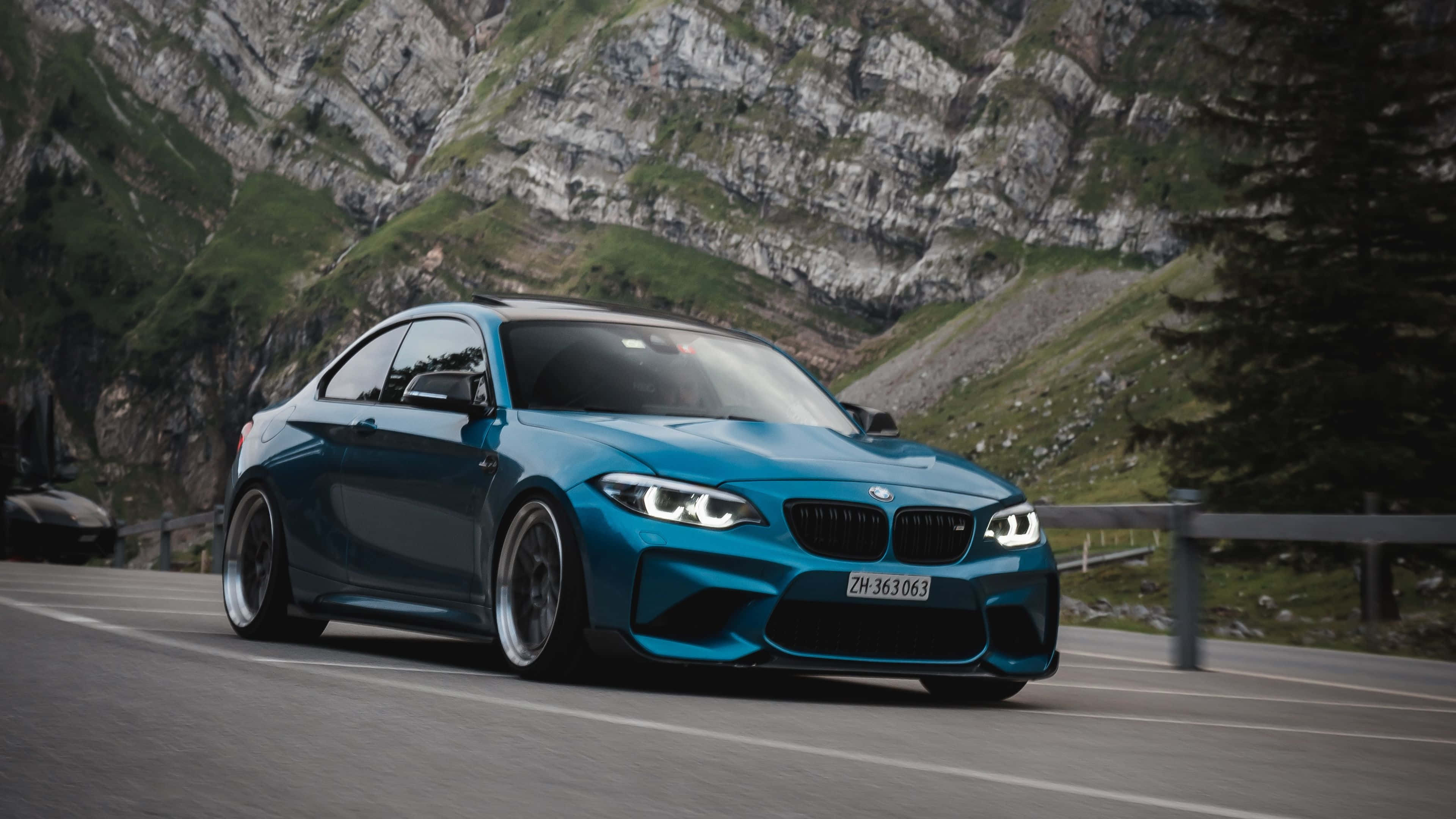 4K BMW 壁紙 桌布 壁紙 — 一輛充滿活力的藍色BMW M2 Coupe在蜿蜒的山路上疾馳，背景是崎嶇的懸崖和綠色的山坡。