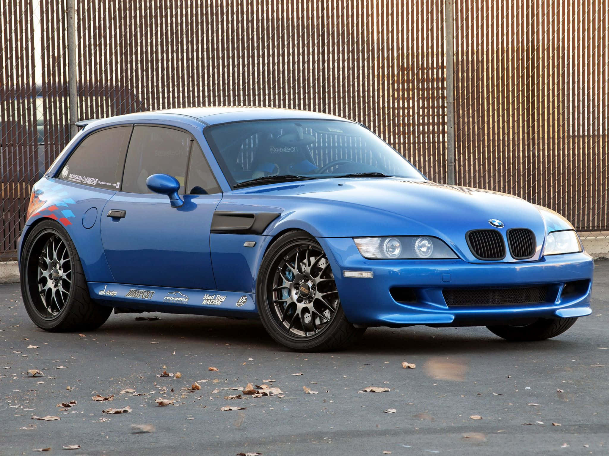 Bmw Z3 桌布 壁紙 — 一輛充滿活力的藍色BMW M3賽車，帶有定制貼花和黑色輪圈，停在木柵欄附近的瀝青路面上。