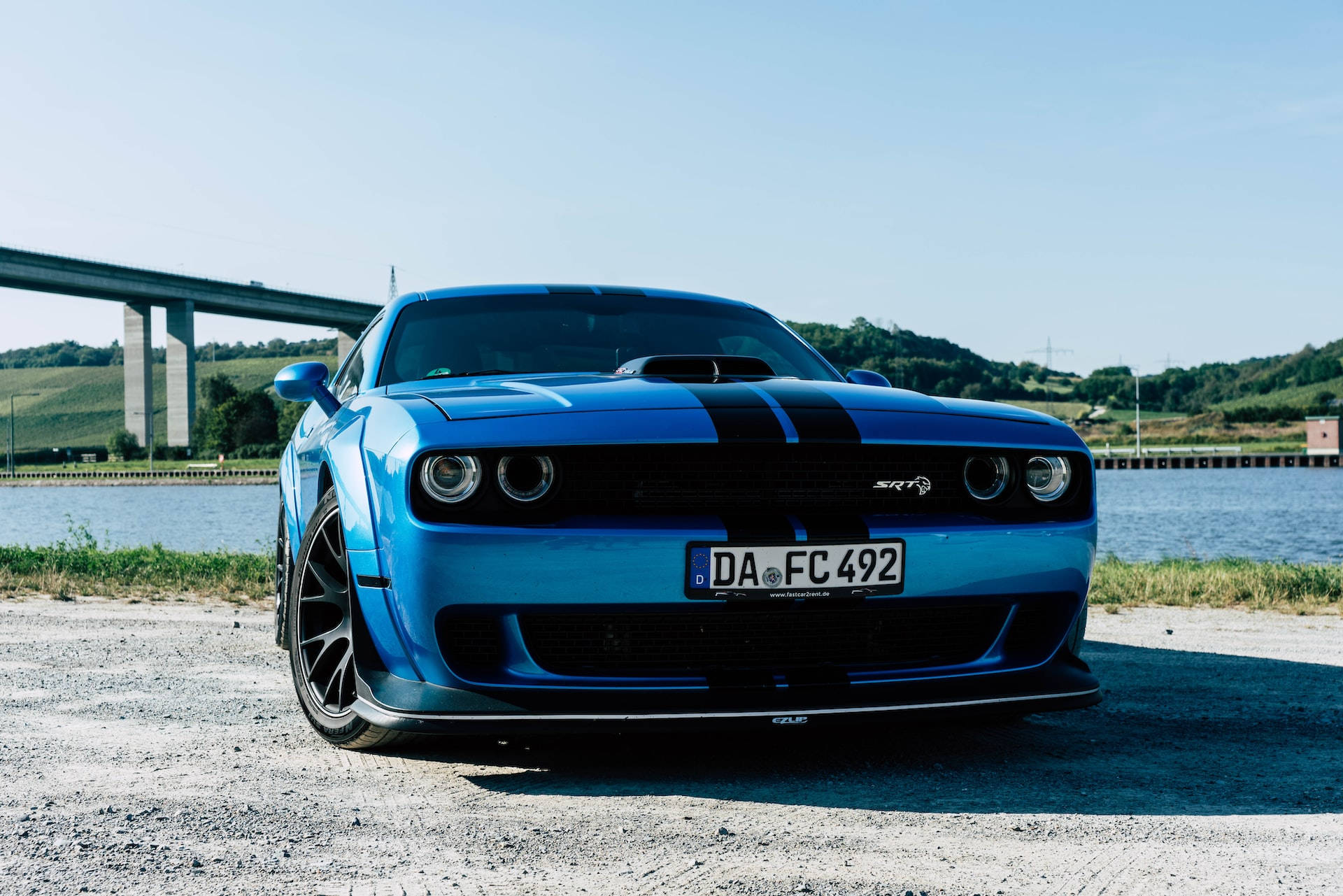 自然 桌布 壁紙 — 一輛充滿活力的藍色Dodge Challenger SRT，帶有黑色賽車條紋，停在晴朗天空下近水的礫石上。