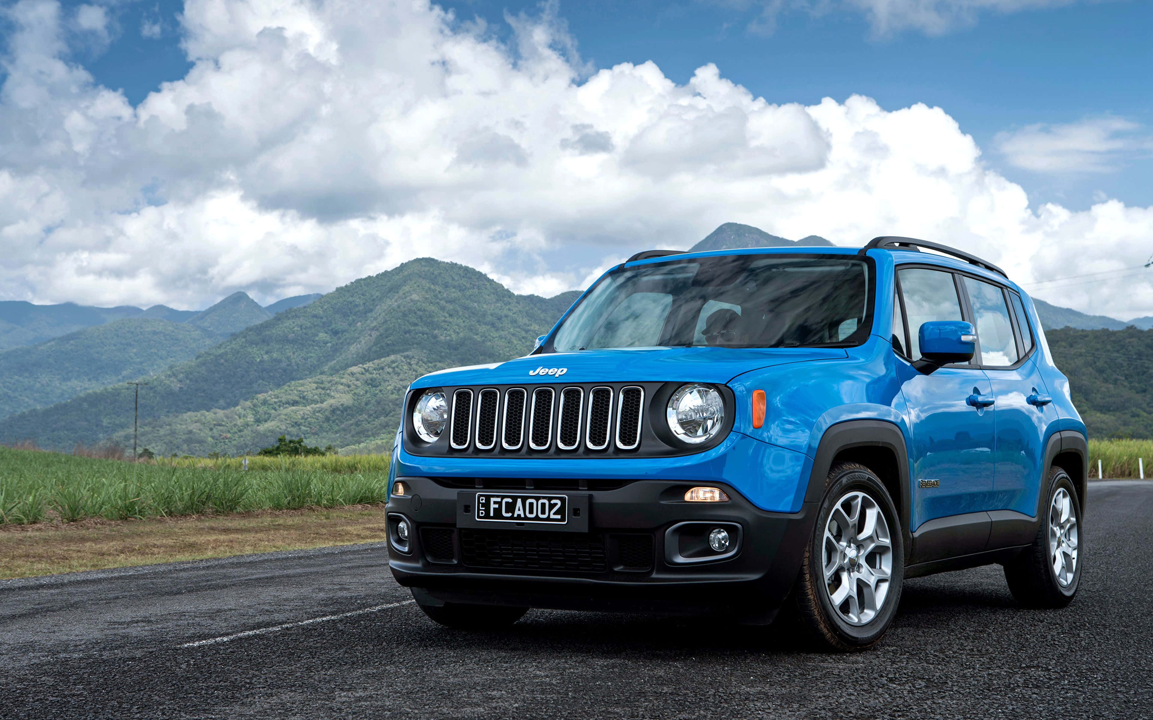 Jeep Renegade 桌布 壁紙 — 一輛充滿活力的藍色Jeep Renegade SUV在鋪砌的道路上行駛，周圍是鬱鬱蔥蔥的綠色山脈和蓬鬆的白雲，在明亮的藍天下。