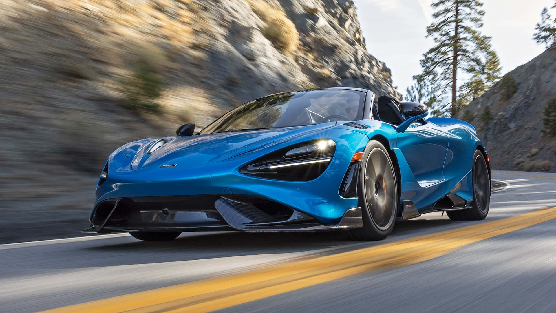 Mclaren 765lt 桌布 壁紙 — 一輛充滿活力的藍色McLaren 720S在蜿蜒的山路上疾馳，背景是模糊的岩石懸崖和松樹。