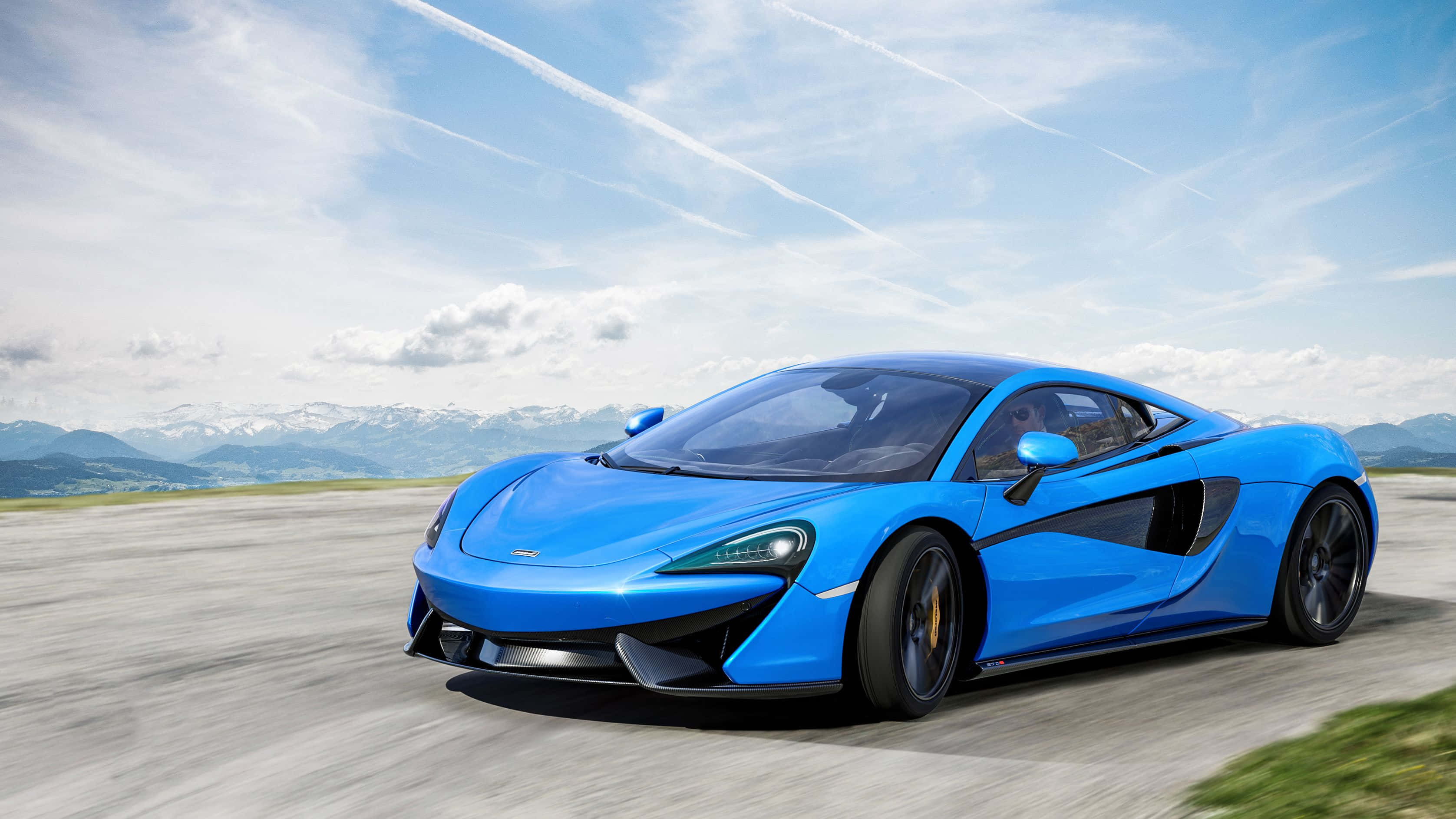 麥拿侖 桌布 壁紙 — 一輛充滿活力的藍色McLaren跑車在明亮的天空下沿著山路飛馳，遠處是白雪皚皚的山峰。