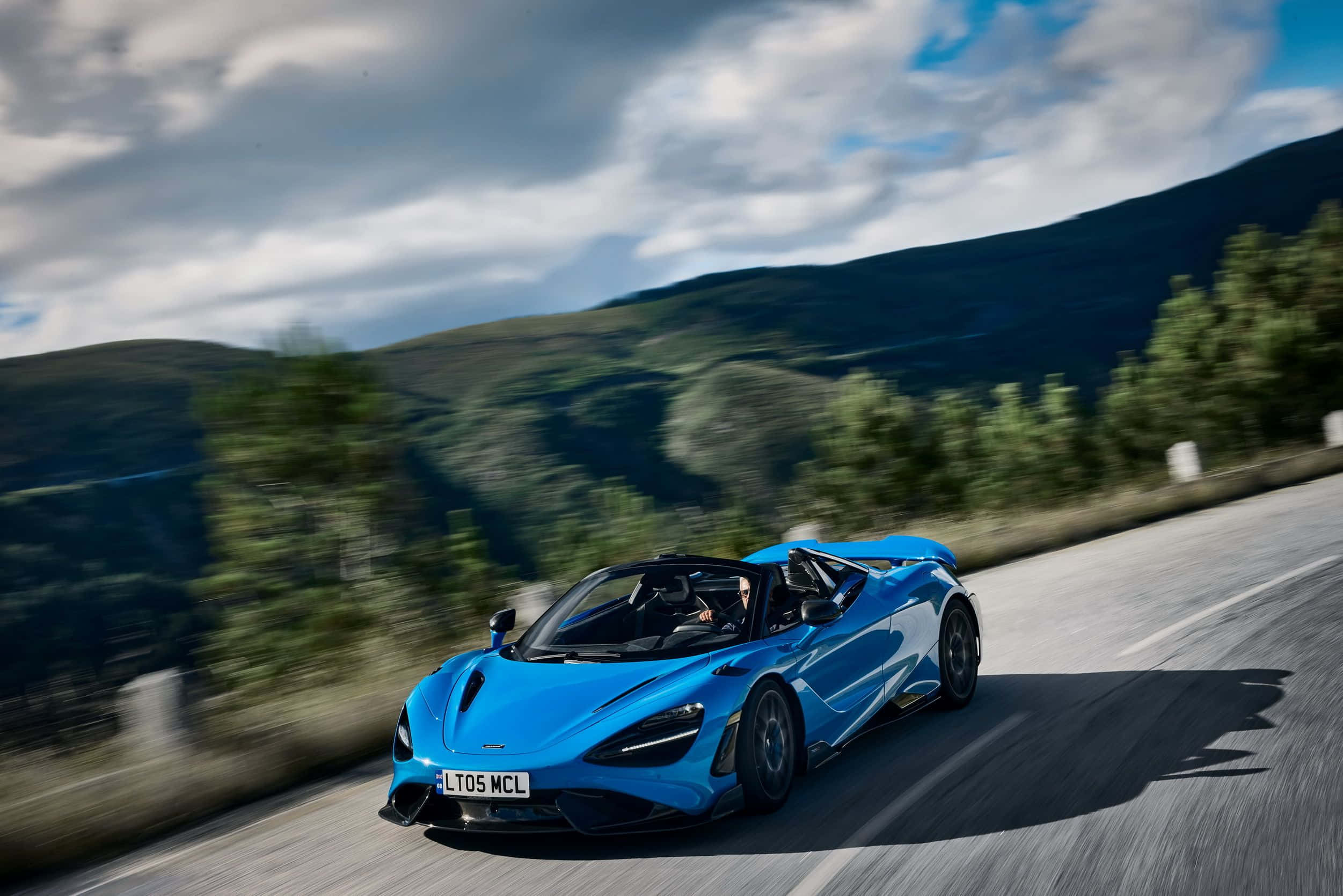 Mclaren 765lt 桌布 壁紙 — 一輛充滿活力的藍色McLaren跑車在部分多雲的天空下沿著蜿蜒的山路飛馳。