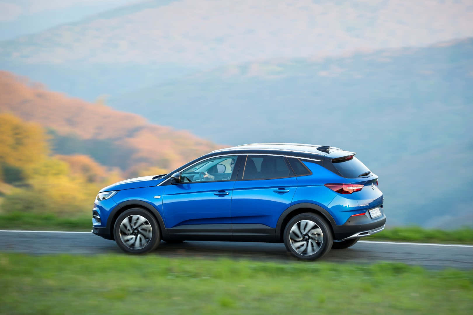 Opel Grandland X 桌布 壁紙 — 一輛充滿活力的藍色SUV沿著蜿蜒的山路行駛，背景是朦朧的秋日山丘。