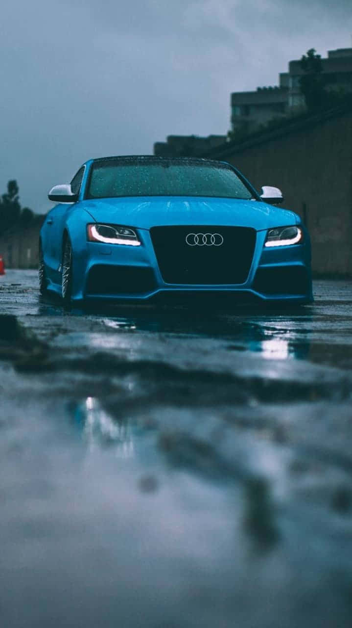  壁紙 — 一輛充滿活力的藍色奧迪跑車在陰天閃閃發光，車頭燈刺破黑暗，濕滑的路面映照出其流線型車身。