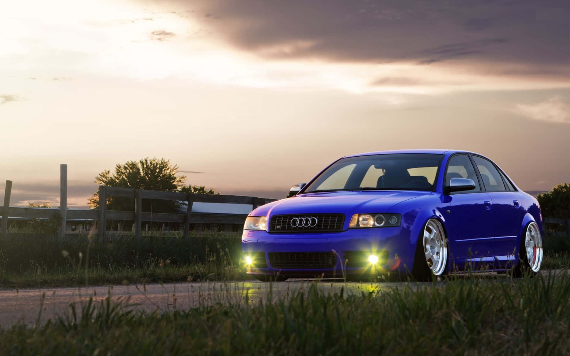 Audi S4 桌布 壁紙 — 一輛充滿活力的藍色奧迪轎車，車頭燈閃爍，停在日落時分的路邊，背景有草地和圍欄。