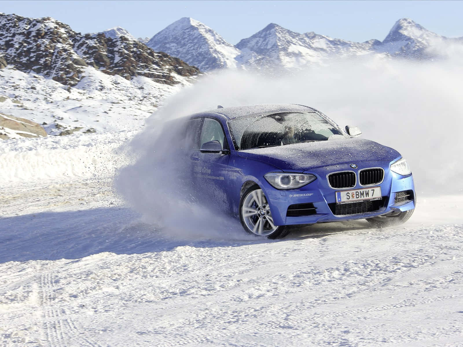Bmw 1 Series 桌布 壁紙 — 一輛充滿活力的藍色寶馬汽車在晴朗的藍天下，在白雪覆蓋的山口疾馳，揚起雪花。