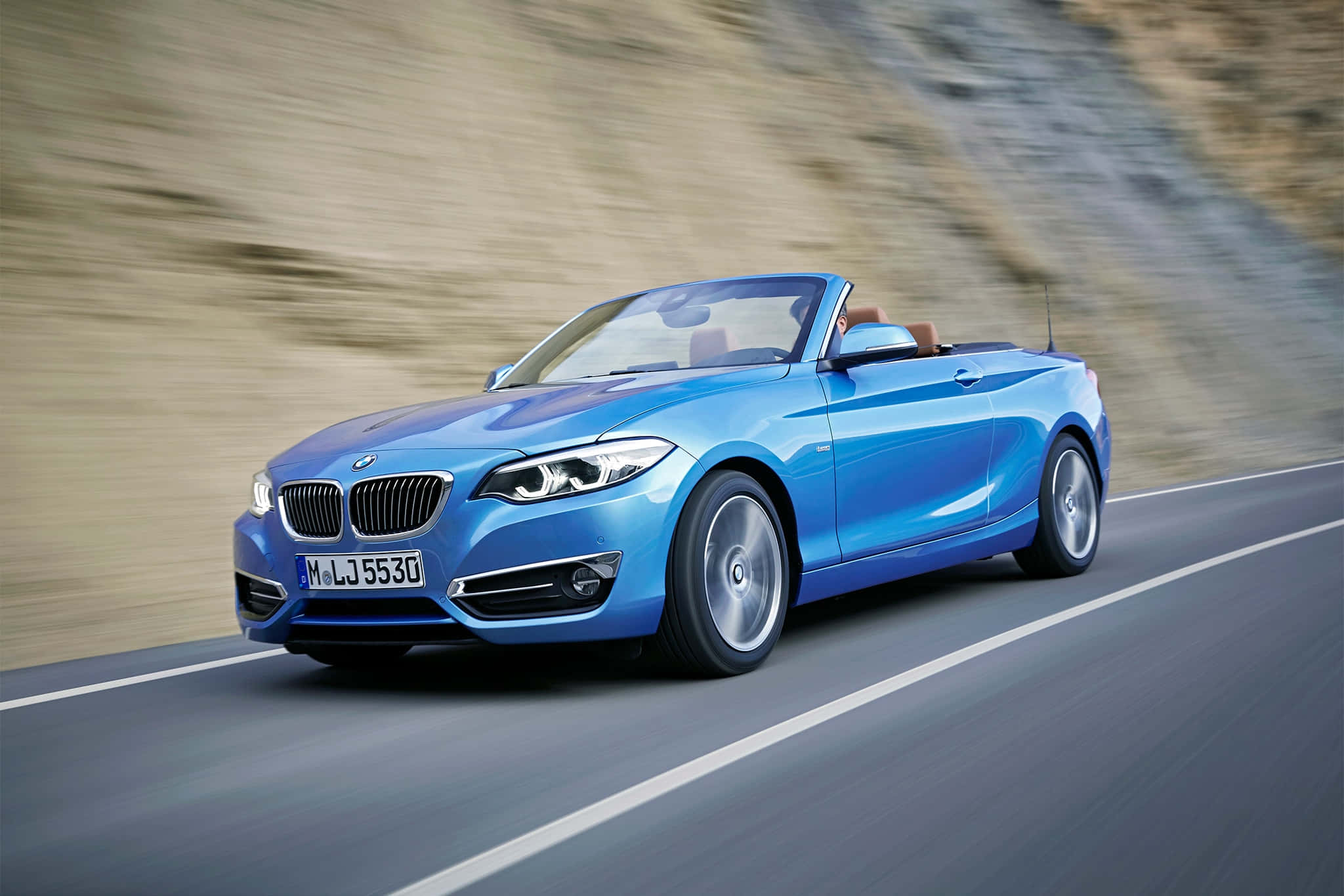 Bmw 2 Series 桌布 壁紙 — 一輛充滿活力的藍色寶馬開篷跑車沿著蜿蜒的道路疾馳，模糊的土色山丘飛掠而過。
