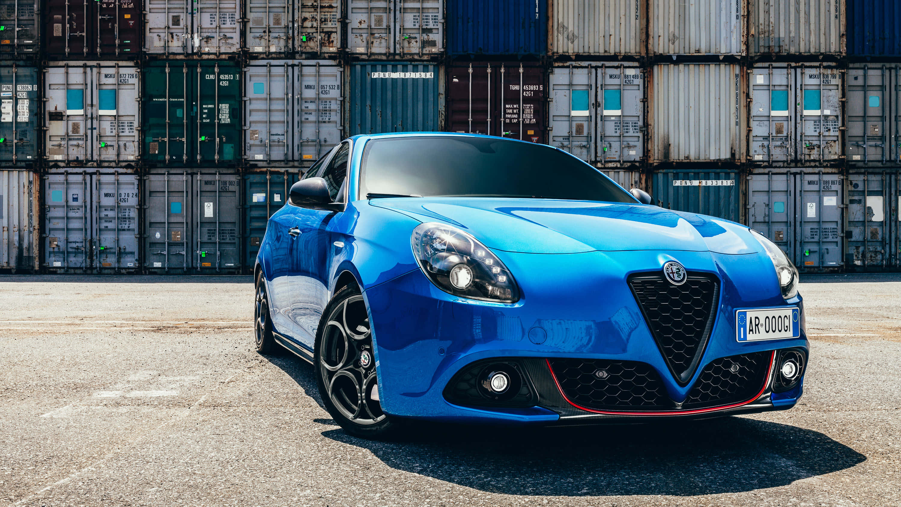 Alfa Romeo Giulietta 桌布 壁紙 — 一輛充滿活力的藍色愛快羅密歐跑車在明亮的陽光下閃閃發光，停泊在一堆高聳的、歷經風霜的貨運集裝箱前。