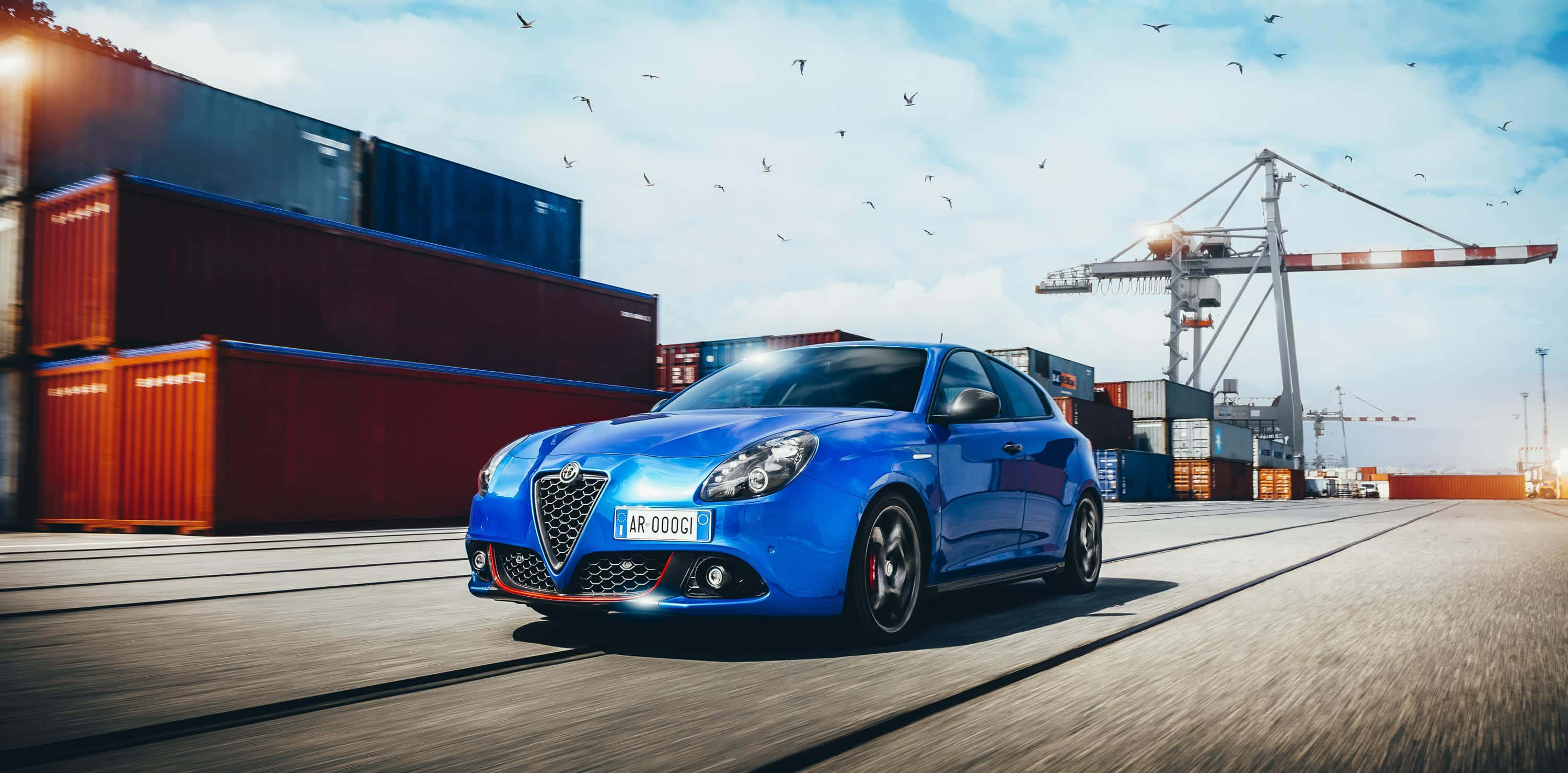 Alfa Romeo Giulietta 桌布 壁紙 — 一輛充滿活力的藍色愛快羅密歐跑車在陽光明媚的港口飛馳，背景是堆疊的貨運集裝箱和起重機。