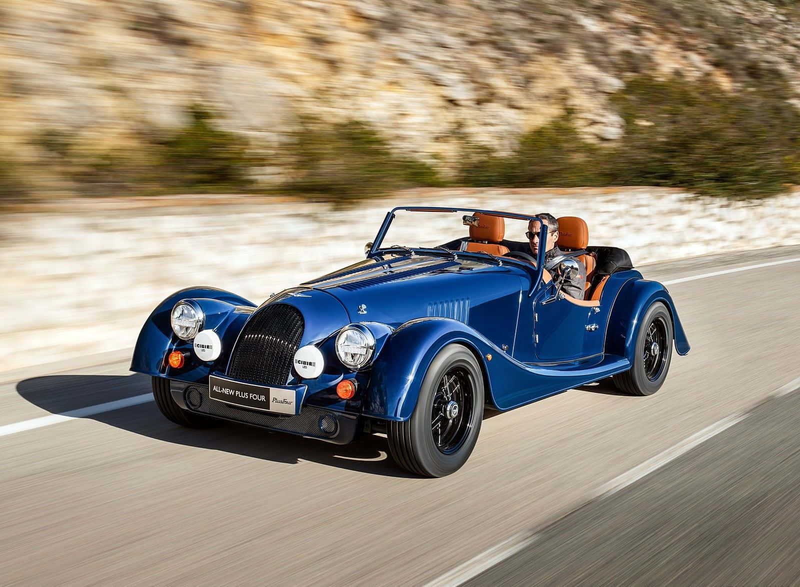 Morgan 桌布 壁紙 — 一輛充滿活力的藍色摩根Plus Four開篷跑車在陽光明媚的山路上飛馳，背景是模糊的岩石懸崖。