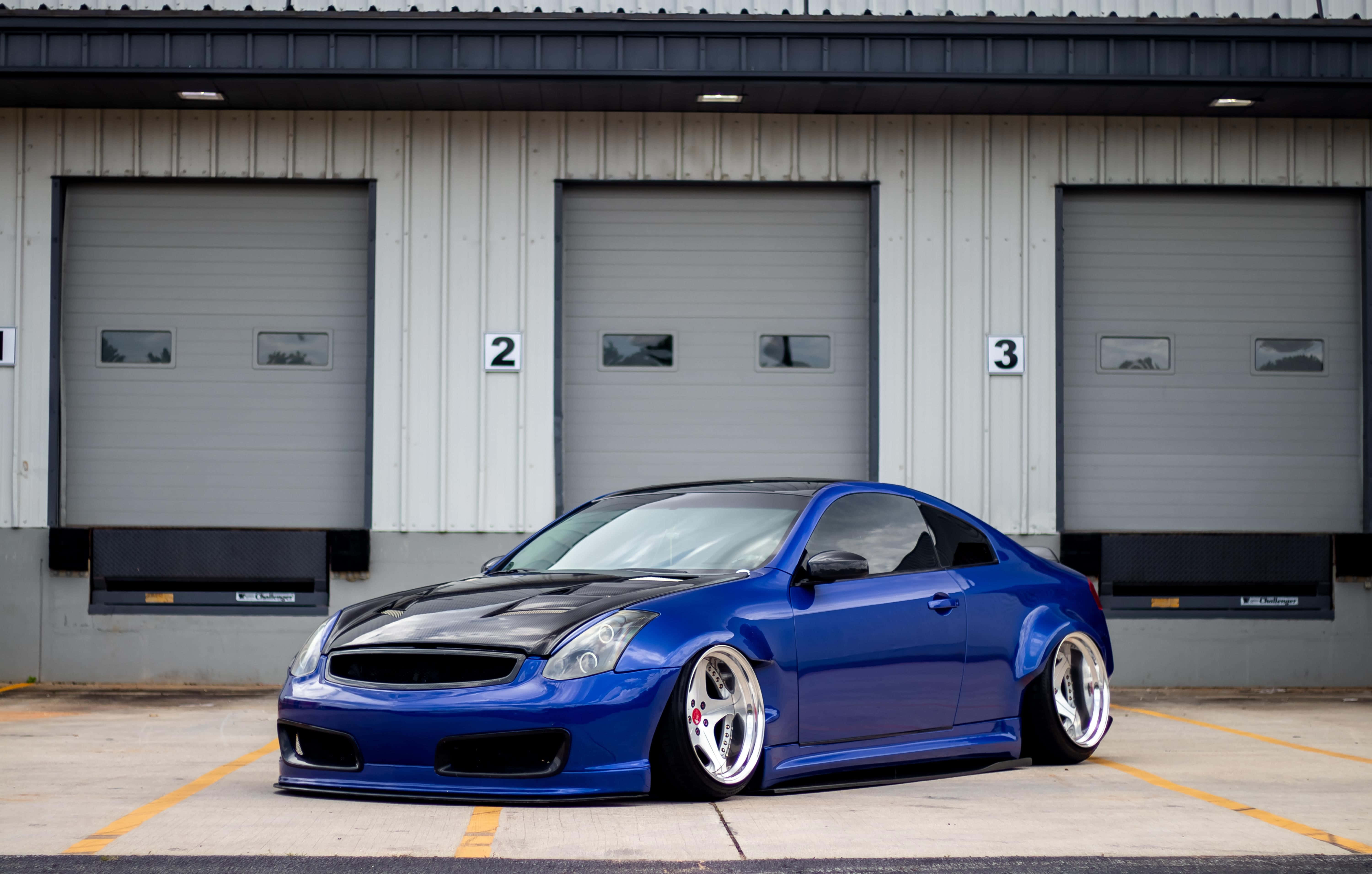Infiniti G35 桌布 壁紙 — 一輛充滿活力的藍色改裝跑車配備定制輪圈，停在一排帶編號的工業車庫門前。