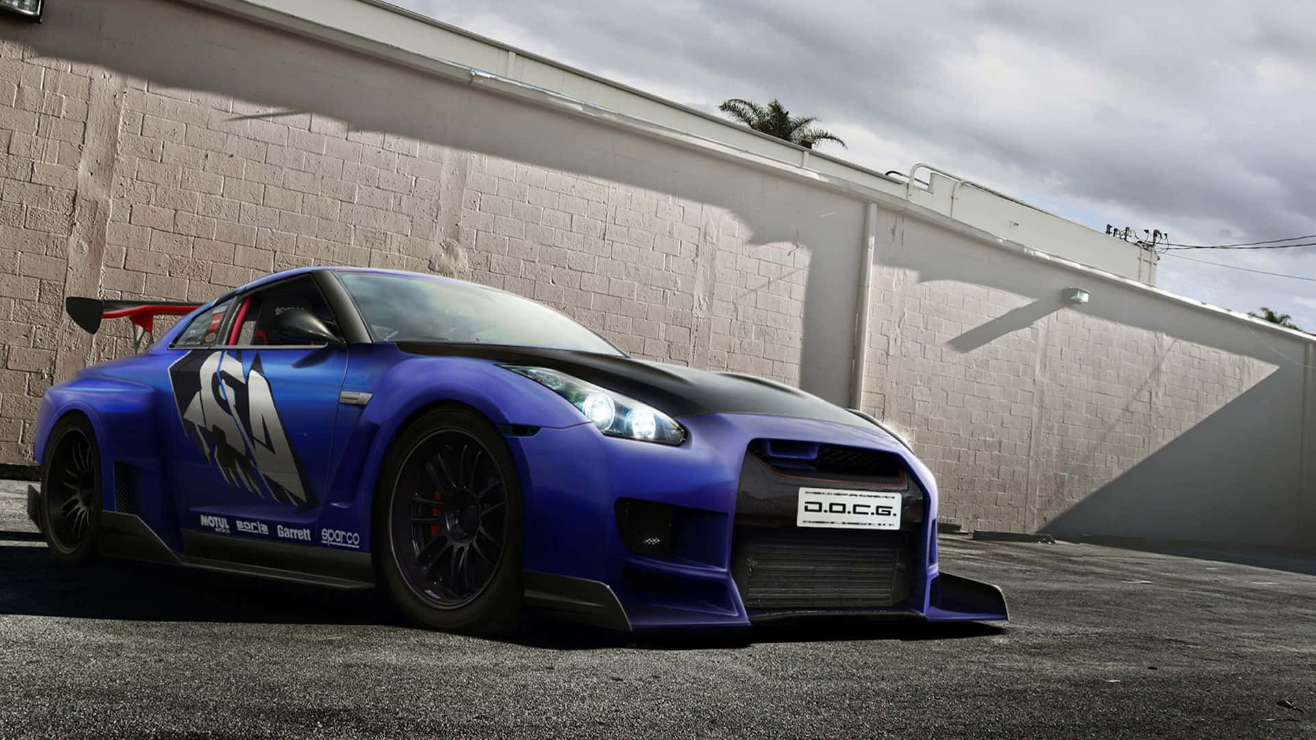 Gtr 桌布 壁紙 — 一輛充滿活力的藍色日產GT-R賽車，帶有黑色飾邊和定制貼花，停在白色磚牆前，天空多雲。