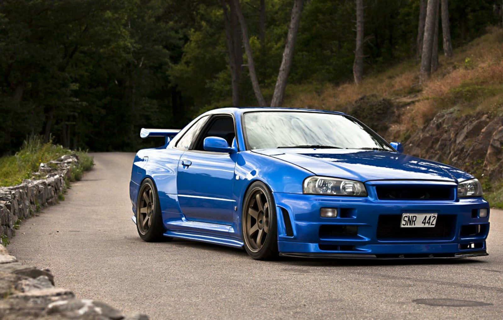 JDM 車 桌布 壁紙 — 一輛充滿活力的藍色日產Skyline GT-R在蜿蜒的林間道路上疾馳，兩旁是石牆和高大的樹木。