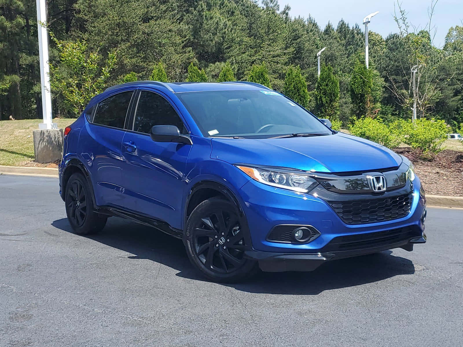 Honda Hr-v 桌布 壁紙 — 一輛充滿活力的藍色本田HR-V，帶有黑色飾邊，停在瀝青路面上，在明亮的日光下，背景有綠樹。