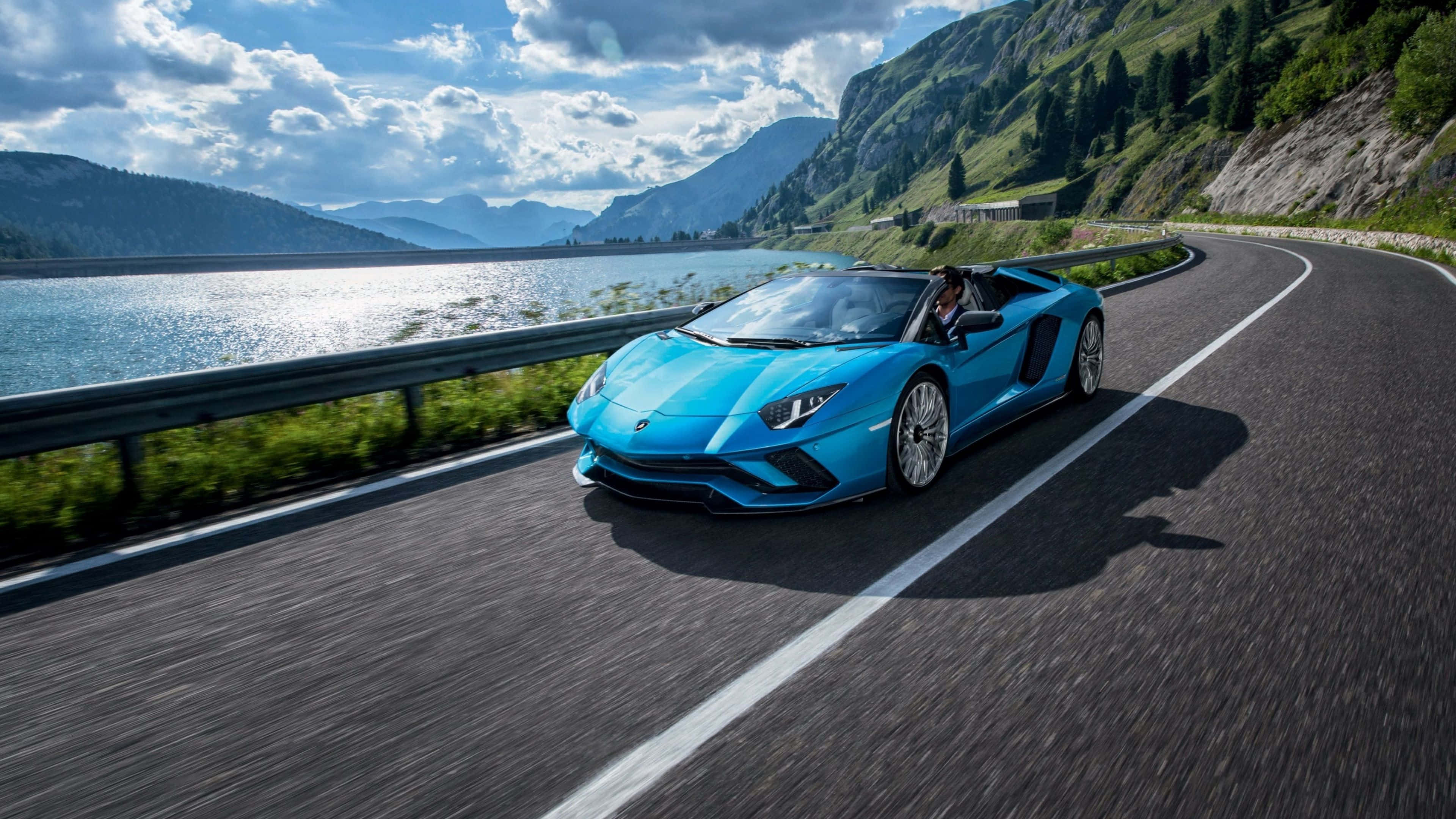 林寶堅尼 4K 桌布 壁紙 — 一輛充滿活力的藍色林寶堅尼 Aventador Roadster 沿著蜿蜒的山路，在波光粼粼的湖邊奔馳，天空部分多雲。