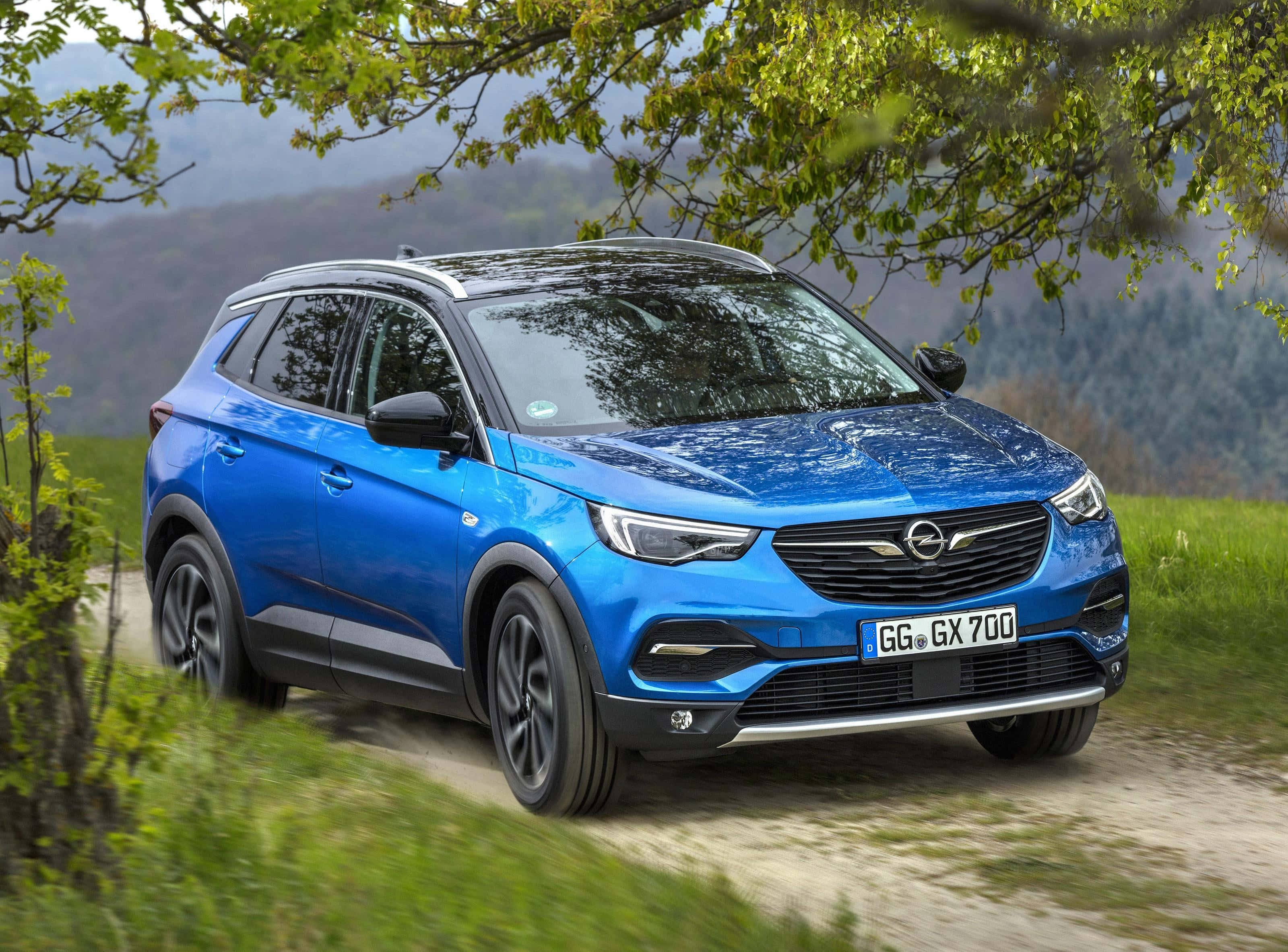 Opel Grandland X 桌布 壁紙 — 一輛充滿活力的藍色歐寶 Grandland X SUV 沿著蜿蜒的泥土路行駛，穿過茂密的綠色森林，背景是山脈。