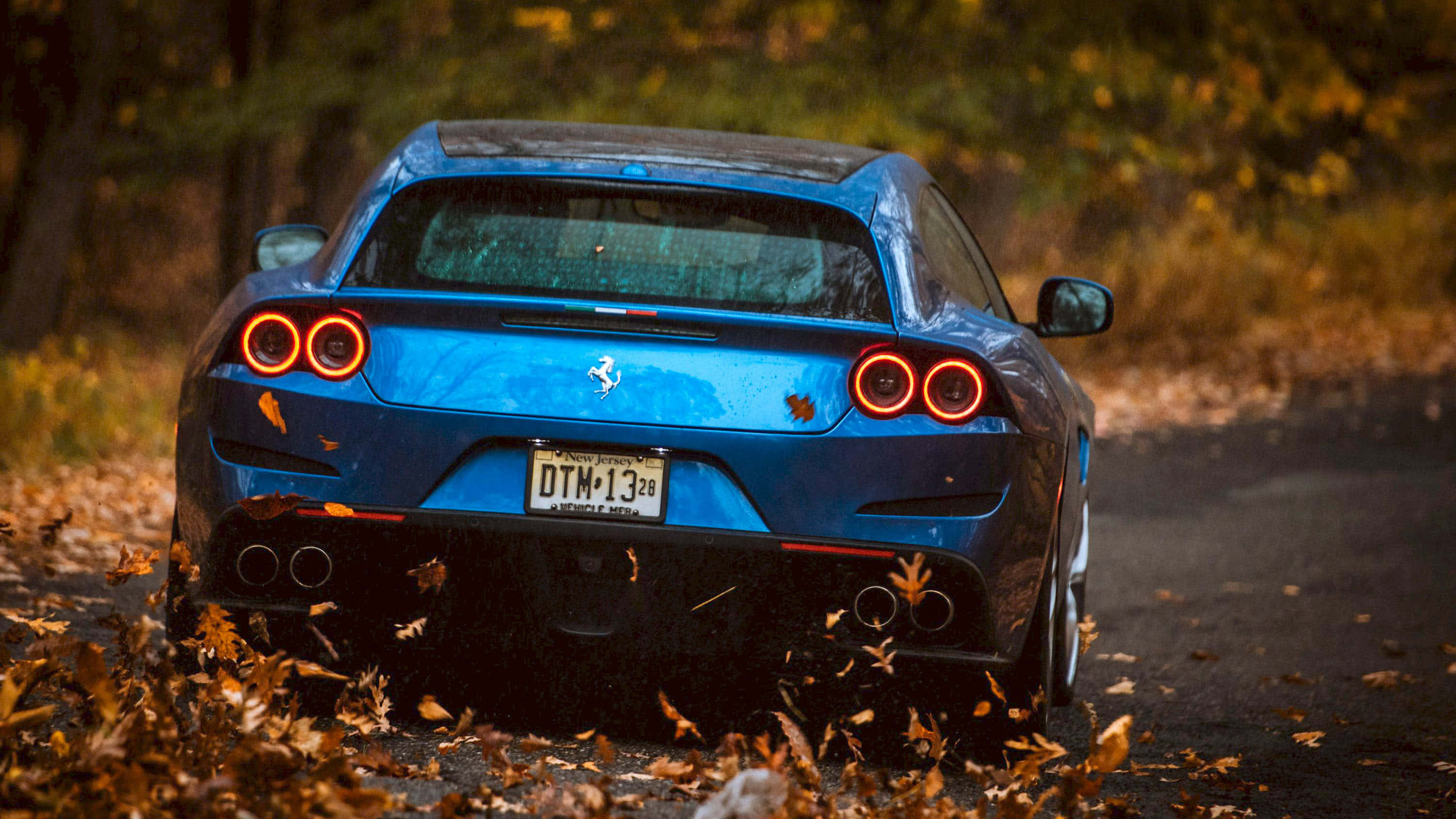Ferrari Gtc4lusso 桌布 壁紙 — 一輛充滿活力的藍色法拉利跑車行駛在鋪滿金色秋葉的森林小徑上。