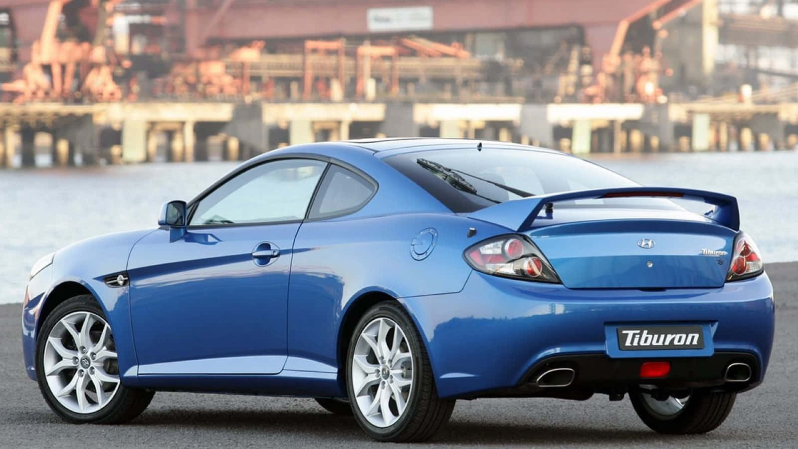 Hyundai Tiburon 桌布 壁紙 — 一輛充滿活力的藍色現代 Tiburon 運動型轎跑車停在水邊，背景有工業起重機。