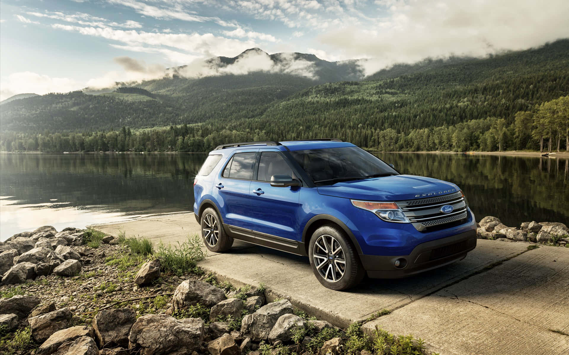 Ford Explorer 桌布 壁紙 — 一輛充滿活力的藍色福特探險者SUV停在湖邊碼頭上，霧氣繚繞的森林山脈在平靜的水面上倒映，天空部分多雲。