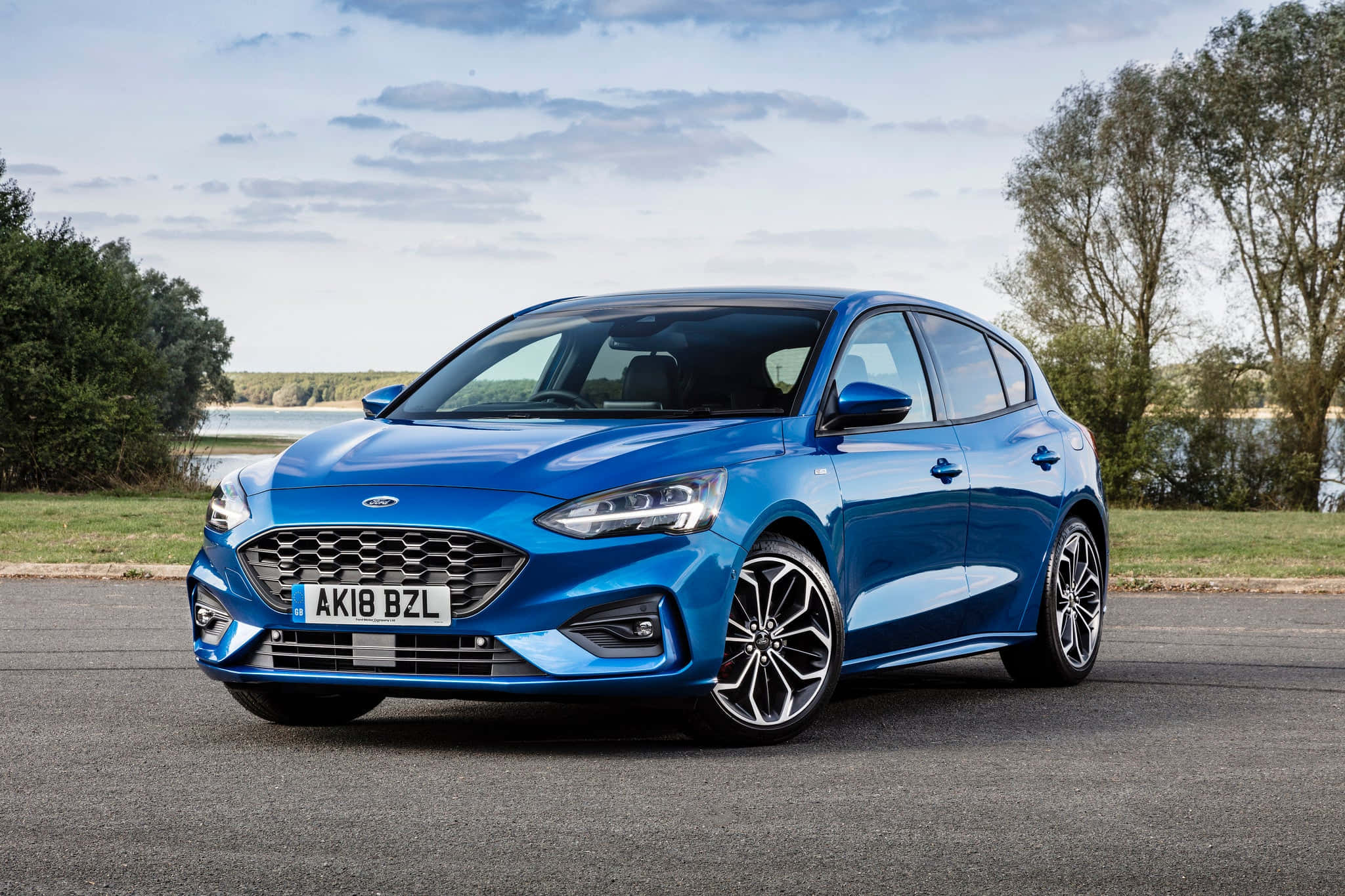 Ford Focus 桌布 壁紙 — 一輛充滿活力的藍色福特福克斯掀背車停在瀝青路上，旁邊是草地、樹木和水域，天空部分多雲。