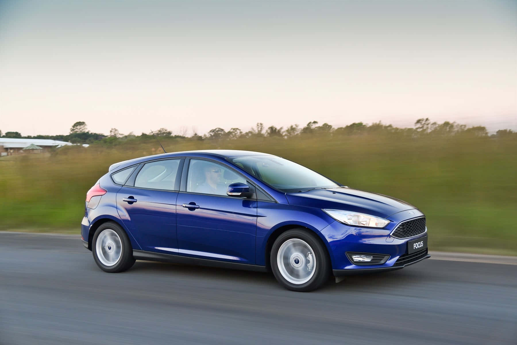 Ford Focus 桌布 壁紙 — 一輛充滿活力的藍色福特福克斯掀背車在鄉村道路上疾馳，背景是模糊的綠意和柔和的日落色調。