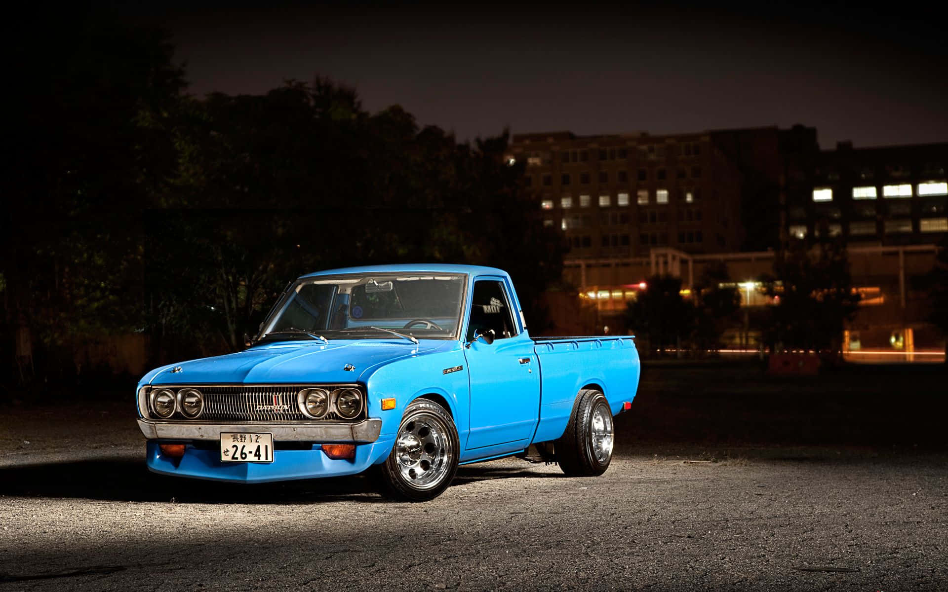 Datsun 桌布 壁紙 — 一輛充滿活力的藍色經典皮卡在夜間聚光燈下閃閃發光，以城市景觀為背景。