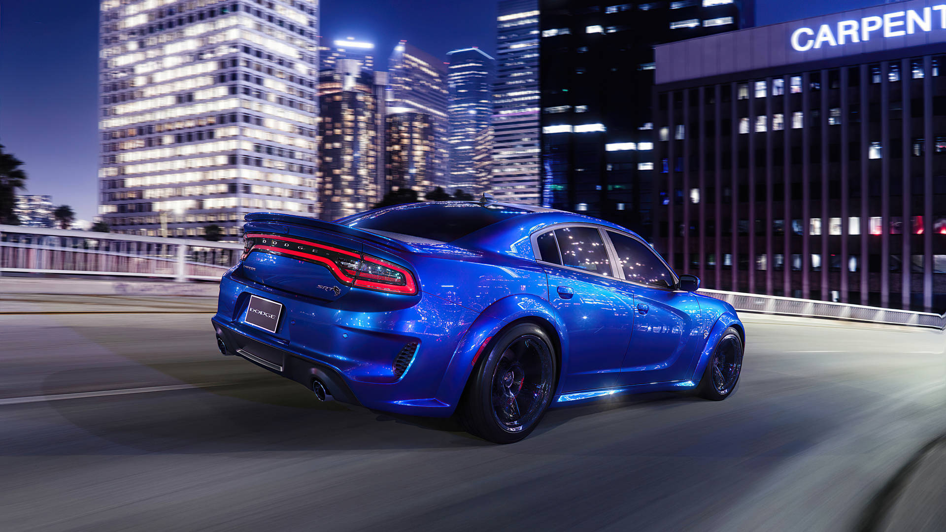 dodge challenger 桌布 壁紙 — 一輛充滿活力的藍色肌肉車在夜間穿過霓虹燈閃爍的城市景觀，背後摩天大樓閃閃發光。
