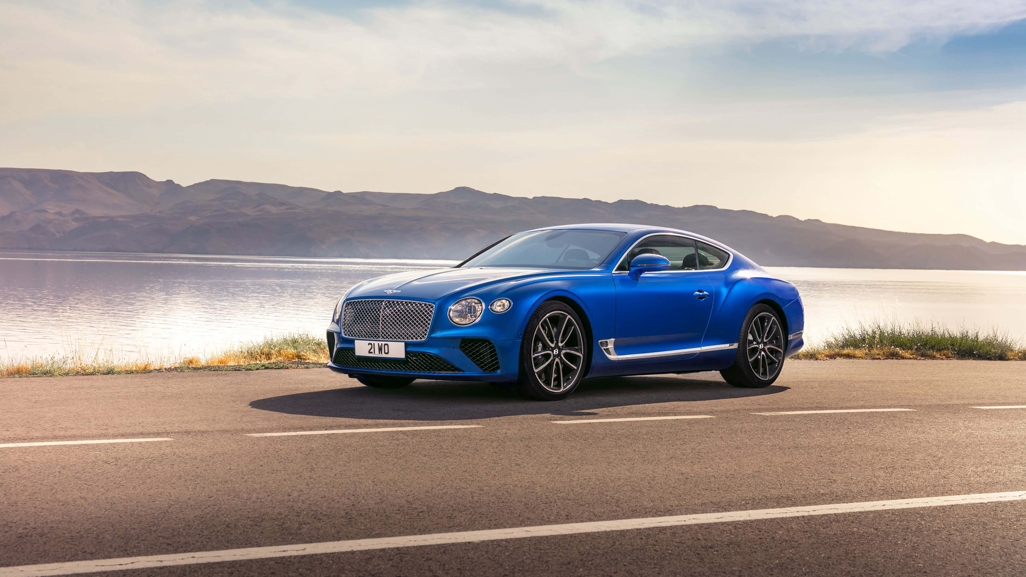 Bentley Continental Gt 桌布 壁紙 — 一輛充滿活力的藍色賓利 Continental GT 雙門跑車停泊在沿海道路上，背景是山脈和寧靜的水面。