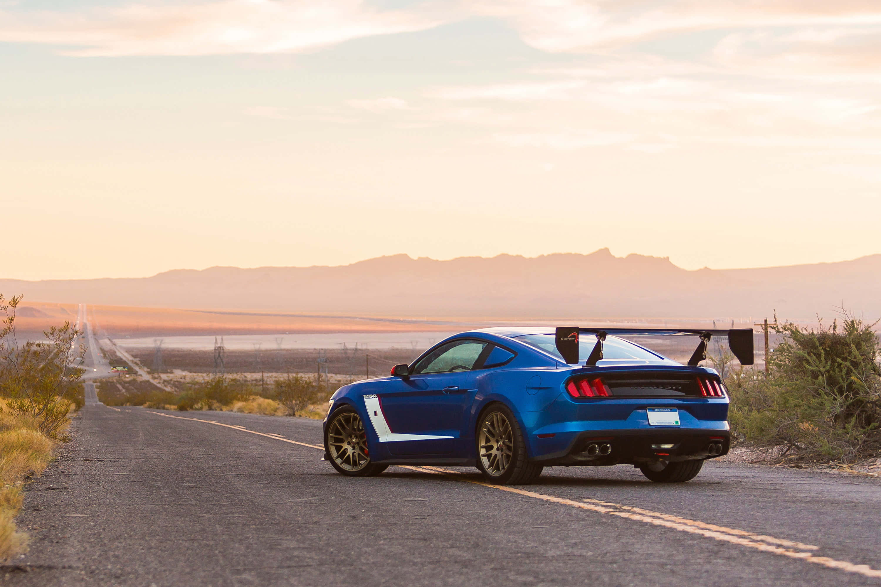 Ford Mustang Roush 桌布 壁紙 — 一輛充滿活力的藍色跑車，帶有大型後擾流板，在日落時分行駛在沙漠高速公路上，背景是遠處的山脈。