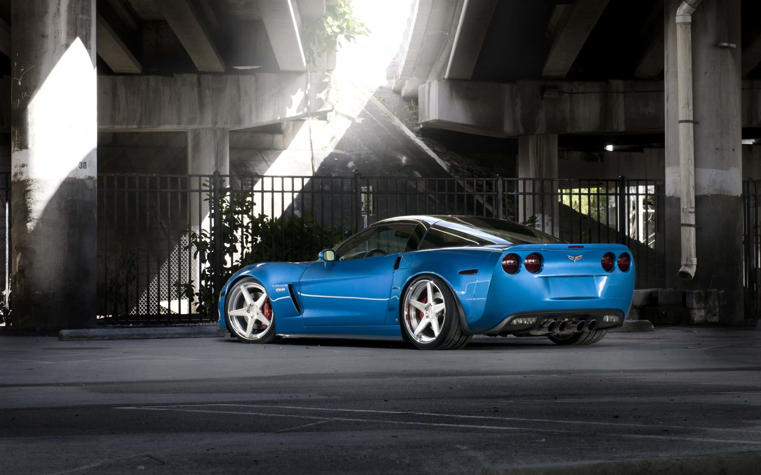 Chevrolet Corvette Z06 桌布 壁紙 — 一輛充滿活力的藍色跑車停泊在城市高架橋下，戲劇性的陽光穿過混凝土樑柱。