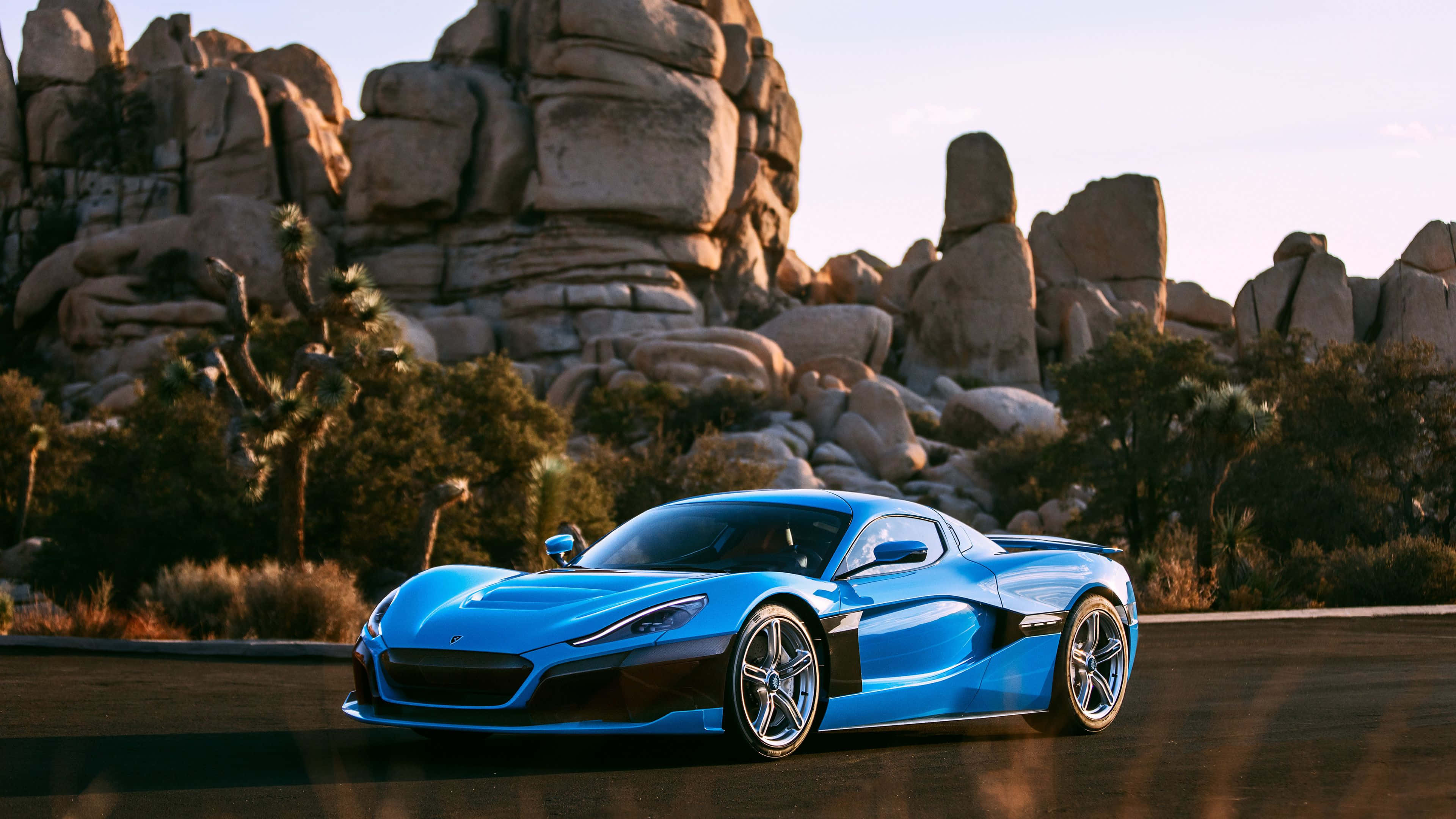 Rimac 桌布 壁紙 — 一輛充滿活力的藍色跑車在沙漠公路上滑行，下方是高聳的岩石 formations，沐浴在金色的日落光中。